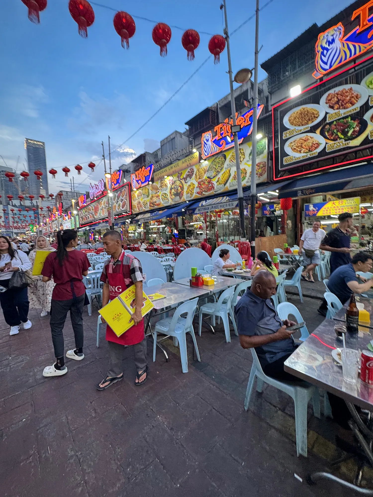Diners at plastic tables under red lanterns on Jalan Alor food street at dusk, Kuala Lumpur.
