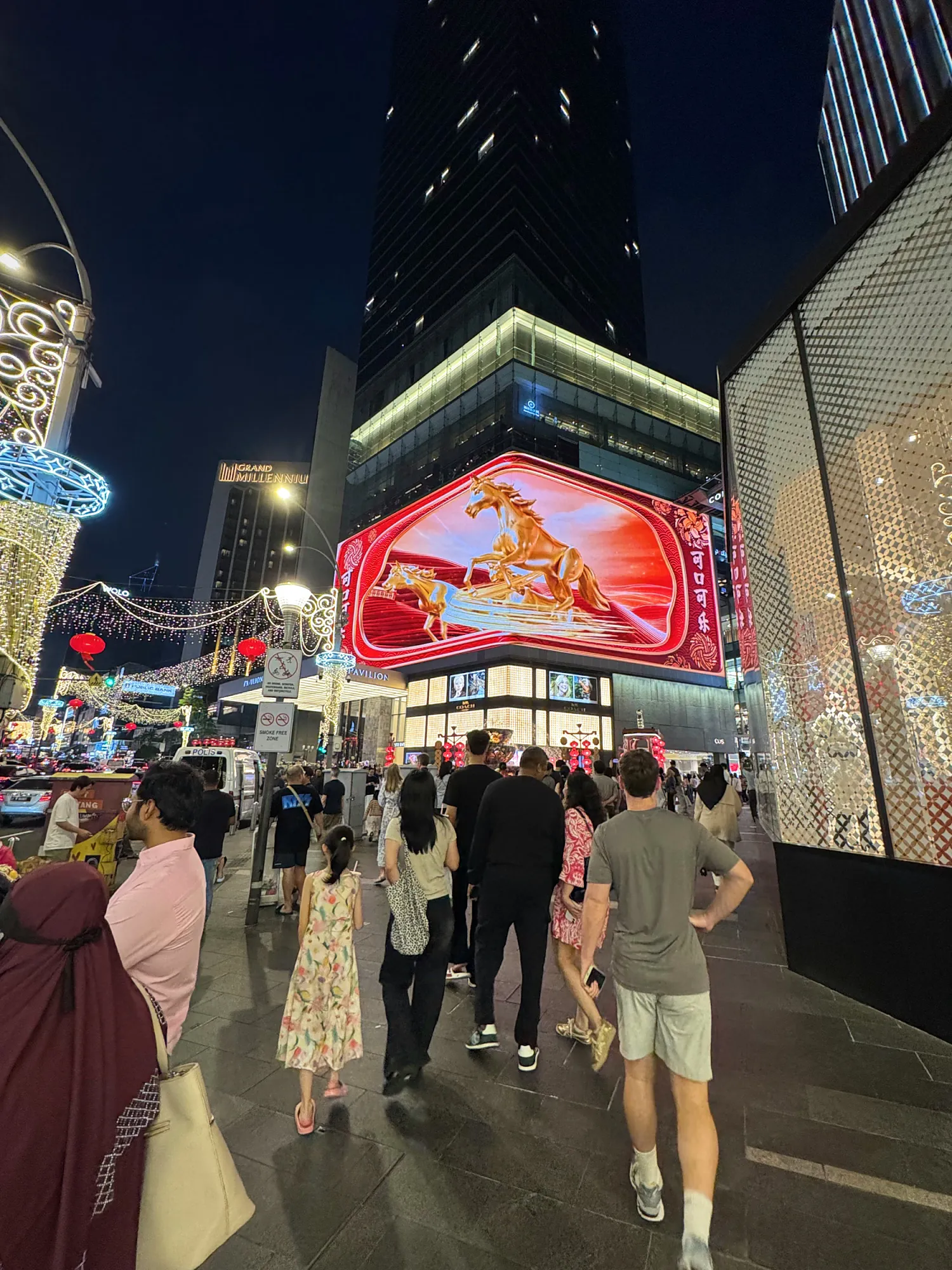 People walking along Bukit Bintang at night past a giant digital billboard showing a golden horse for the Year of the Horse in Kuala Lumpur.