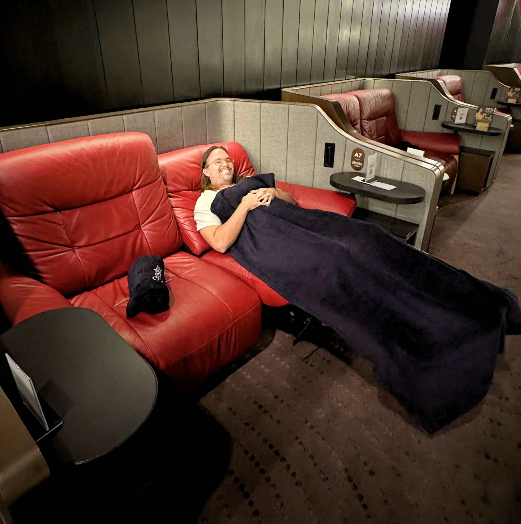Cream relaxing under a navy blanket in a red leather Stressless recliner in a Maybank Comfort Cabin at Aurum Theatre, The Exchange TRX.