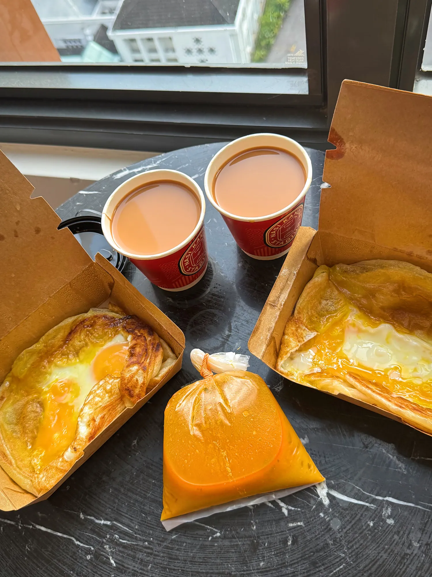Two roti sarang burung in takeaway boxes, each with a runny egg yolk in the middle, beside two cups of teh tarik and a bag of dhal