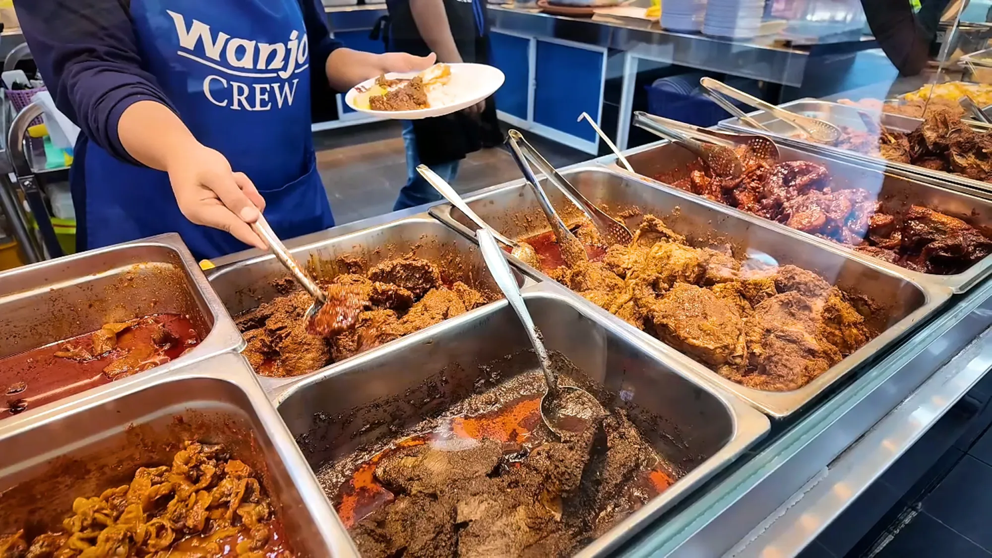 A Wanjo staff member in a blue apron filling a plate from metal trays of rendang, fried chicken, and other side dishes at the counter.