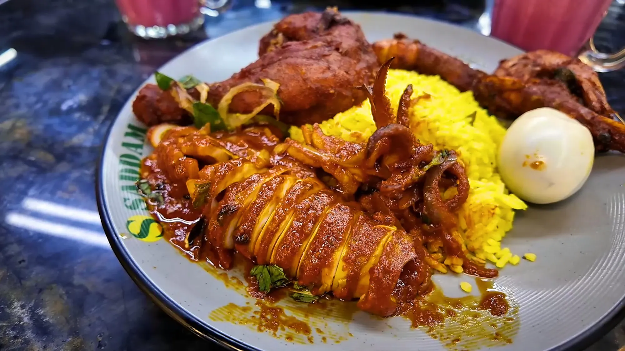 A plate of nasi kandar at Saddam Nasi Kandar in Kuala Lumpur with egg-filled squid in red curry, fried chicken, yellow rice, and a boiled egg.