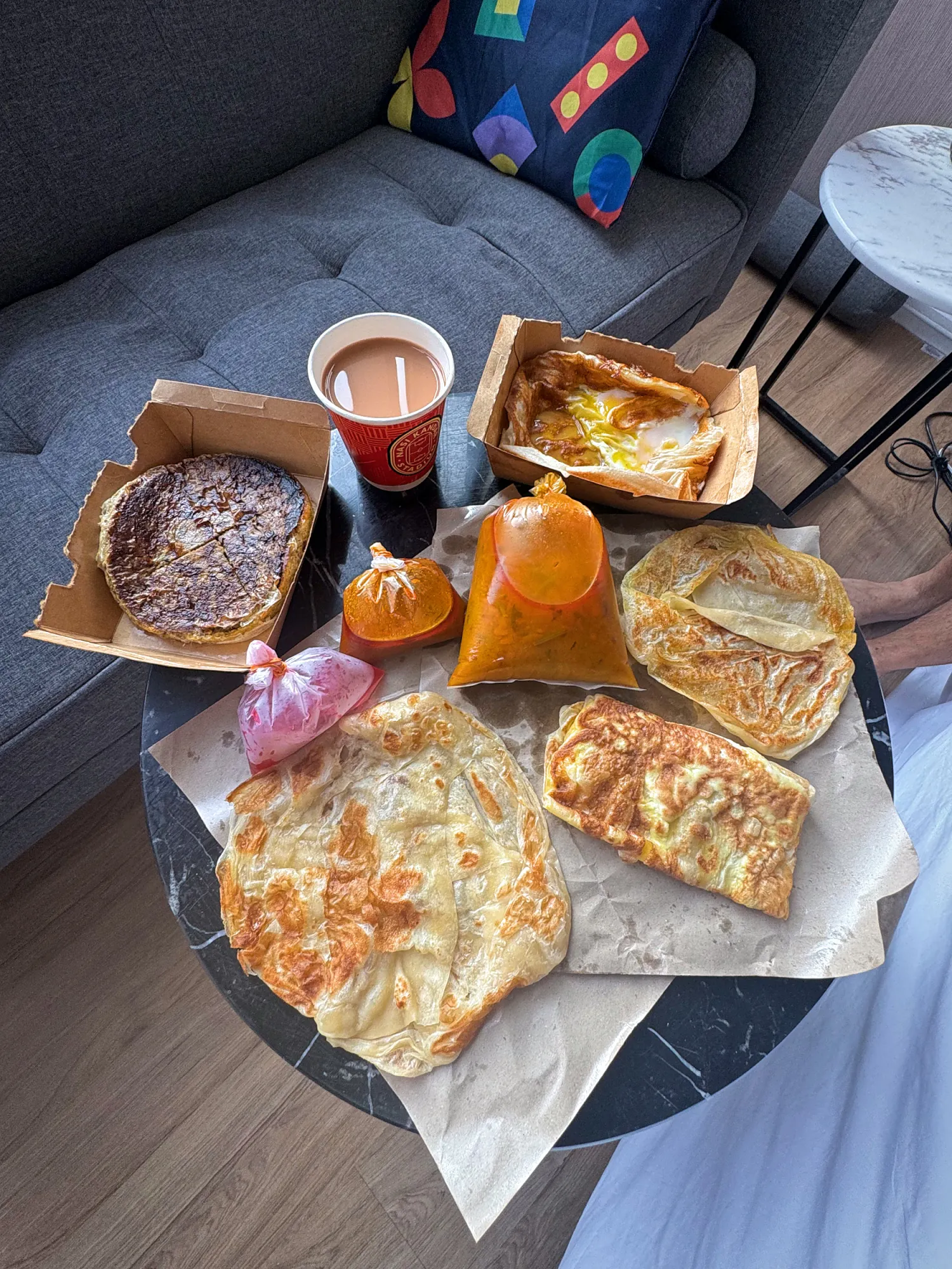 A delivered breakfast of roti canai, murtabak, curries in plastic bags, and a cup of teh tarik on a coffee table in Kuala Lumpur.