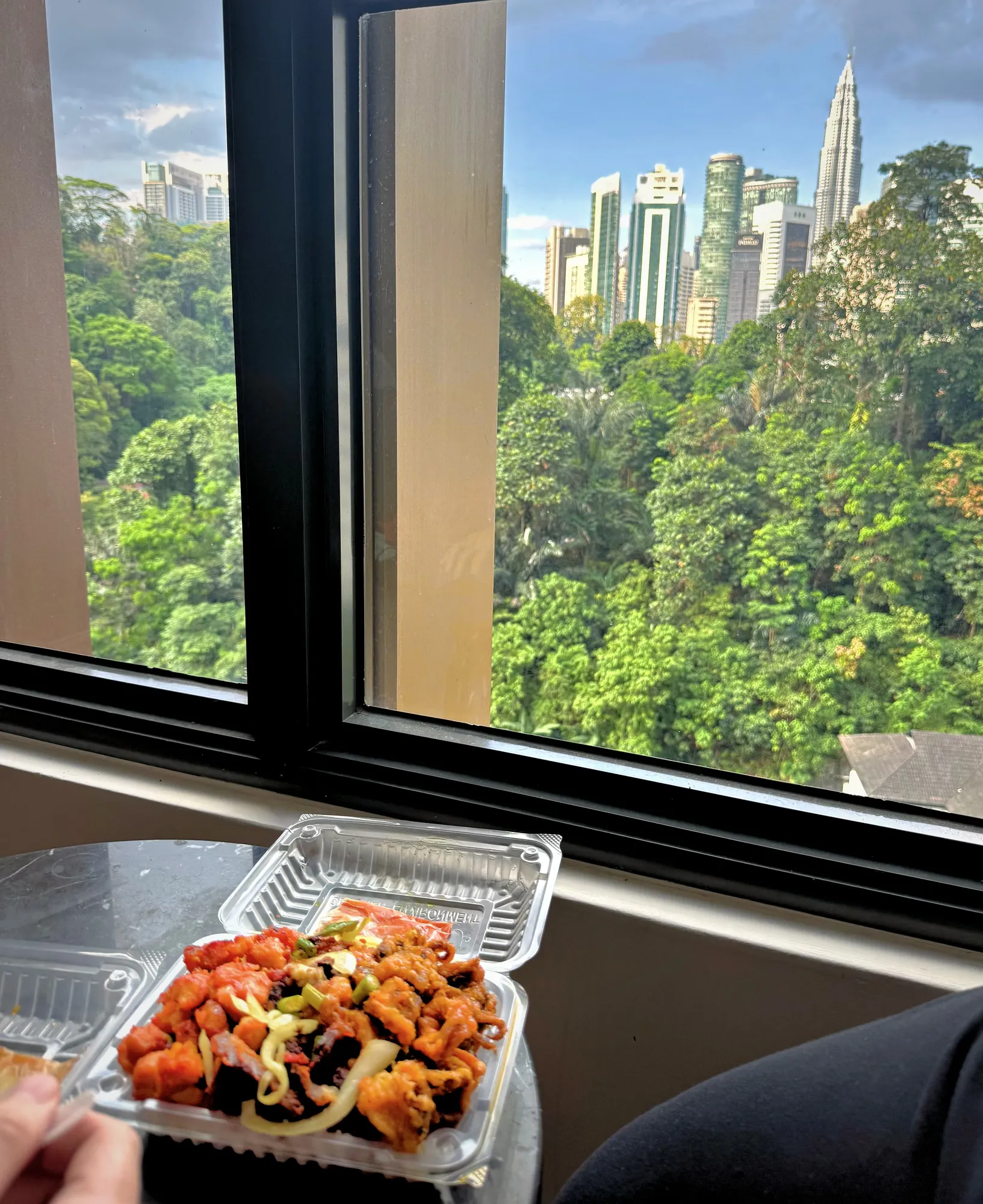 A takeaway box of spicy fried chicken and squid on a table by a window overlooking green treetops and the Kuala Lumpur towers