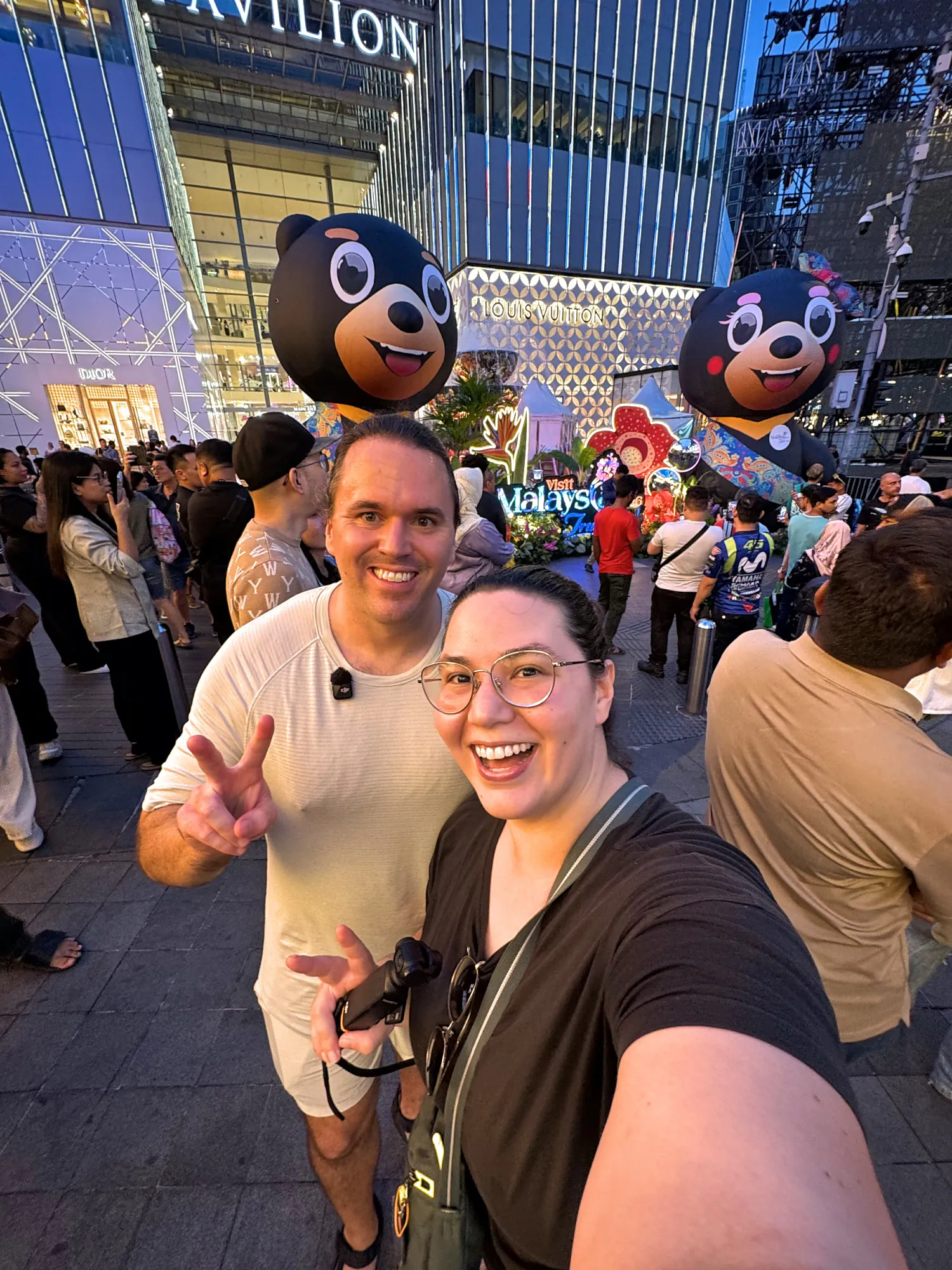 Bernie and Cream taking an evening selfie in the crowd outside Pavilion Kuala Lumpur, with giant Visit Malaysia sun bear figures behind them.
