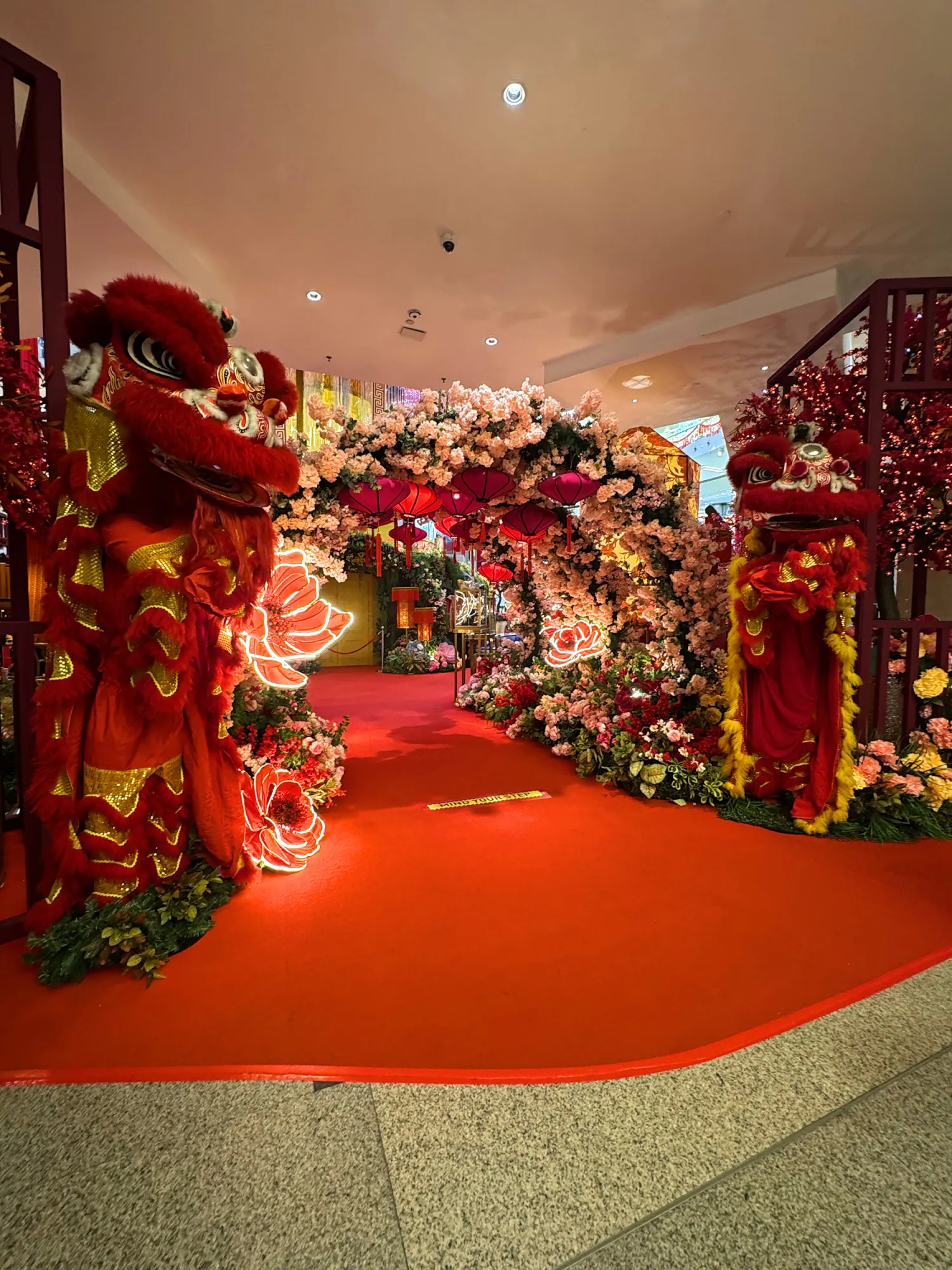 Two red lion dance costumes standing at a Lunar New Year archway covered in cherry blossoms and red lanterns at a mall in Kuala Lumpur.