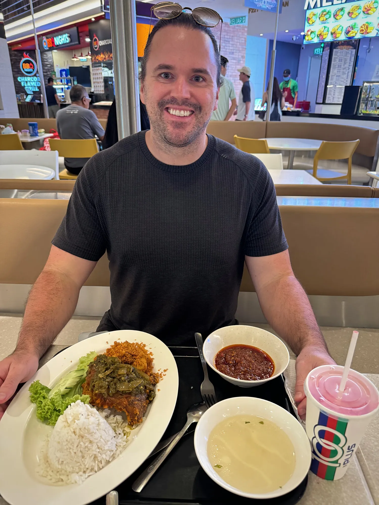 A smiling man at a food court table with a plate of rice, curry and sides and an iced drink in Kuala Lumpur.