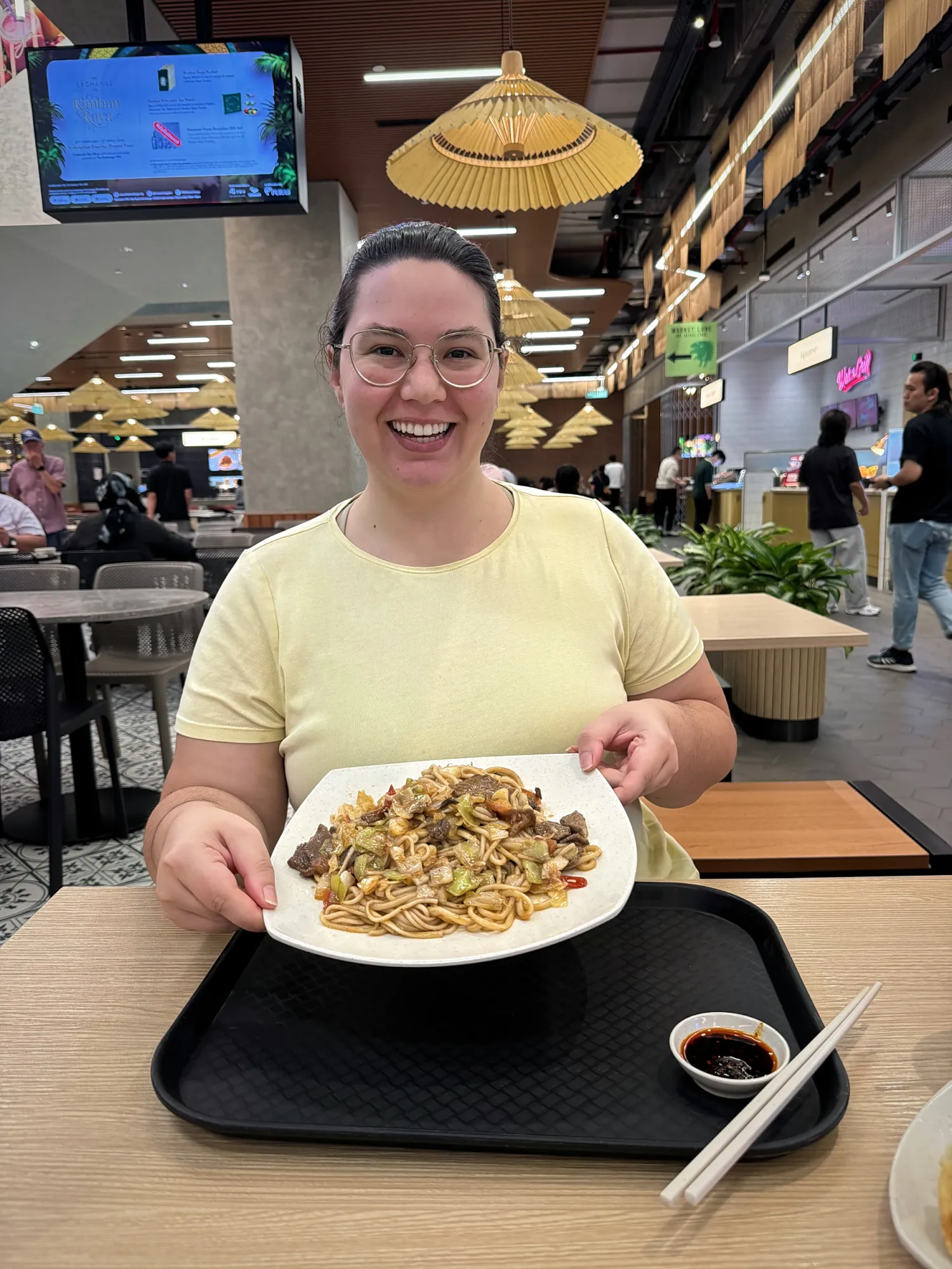 A smiling woman holding a plate of noodles at a table in a Kuala Lumpur mall food court.