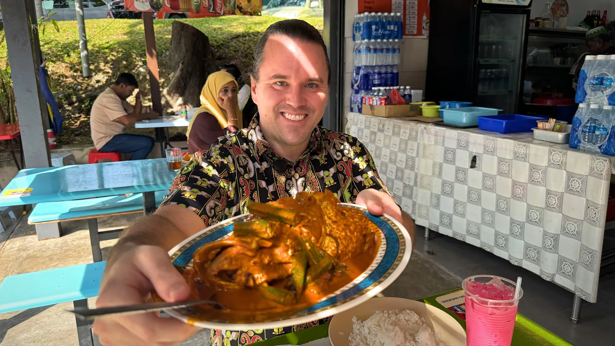Cream smiling and holding up a platter of fish head curry with okra at an outdoor table at Anuar Curry Fish Head in Bangsar, Kuala Lumpur.