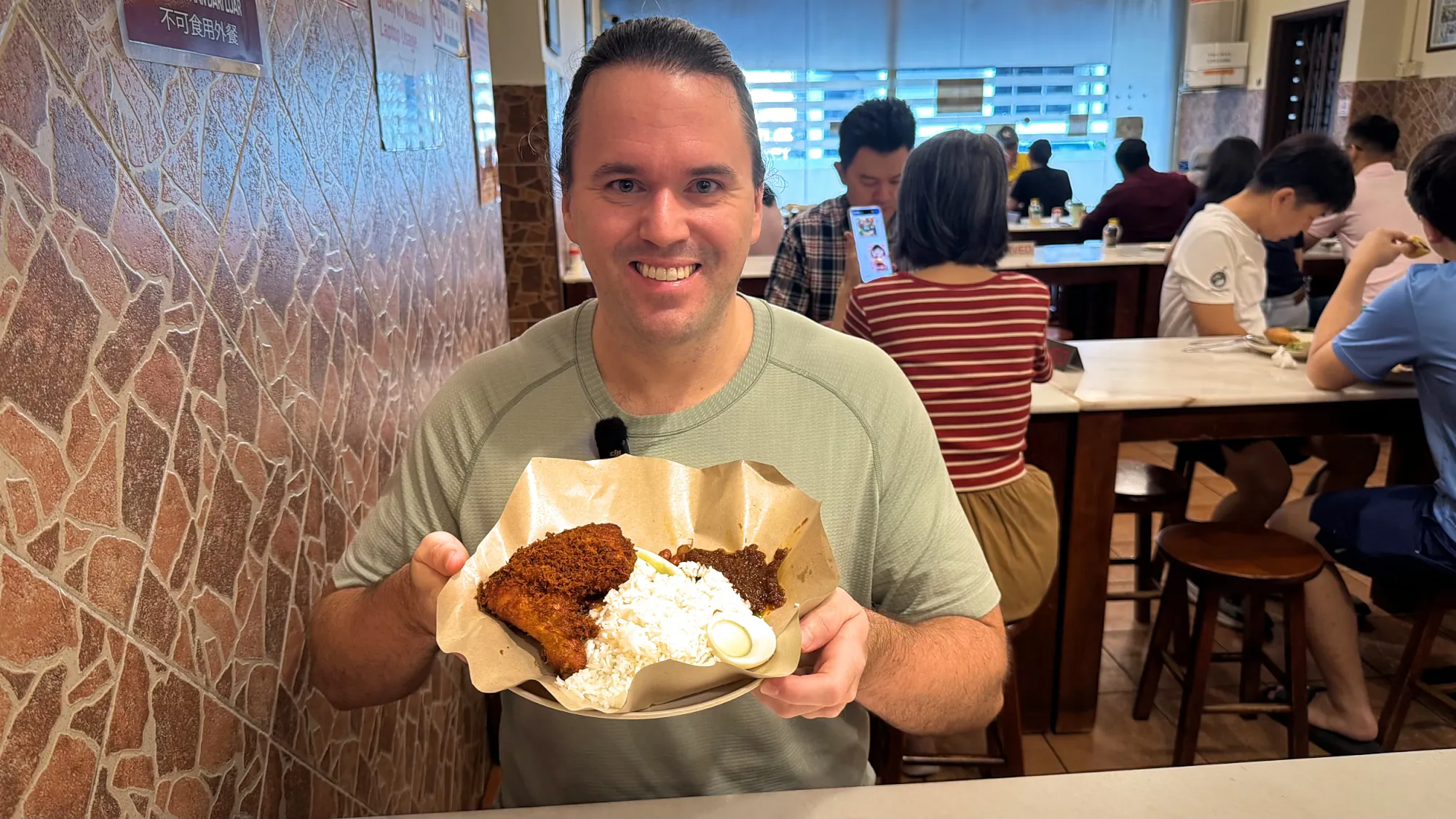 A smiling man holding a plate of nasi lemak with fried chicken inside a restaurant.