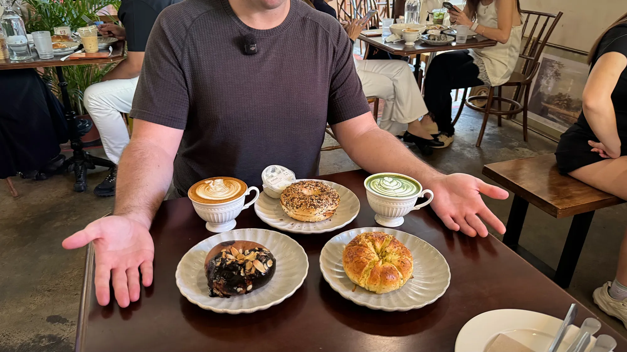 Man at a cafe table gesturing at three bagels, a cappuccino, and a matcha latte at Grumpy Bagels in Kuala Lumpur.