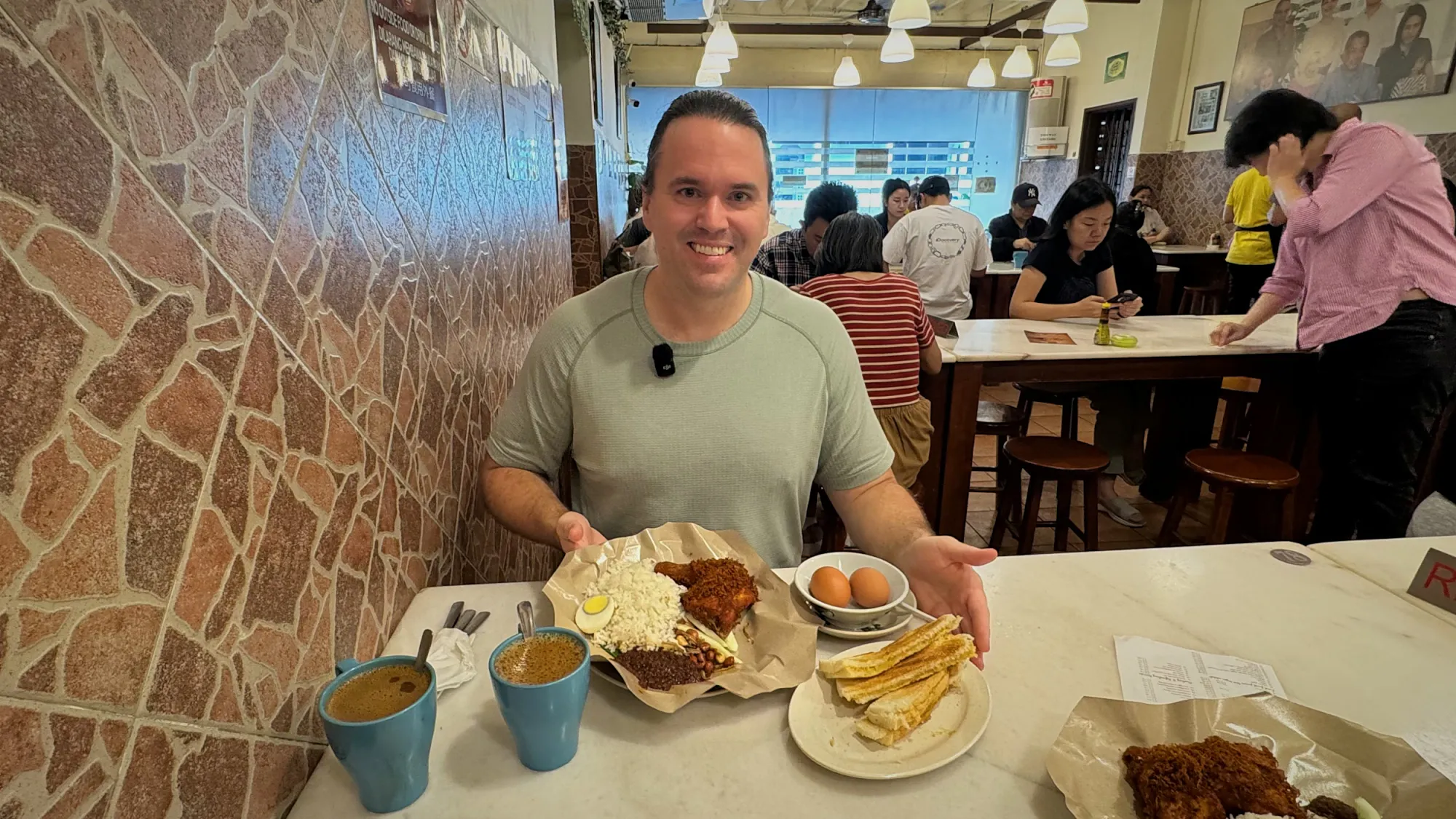 A smiling man presents a plate of nasi lemak with fried chicken and boiled eggs at a restaurant table.