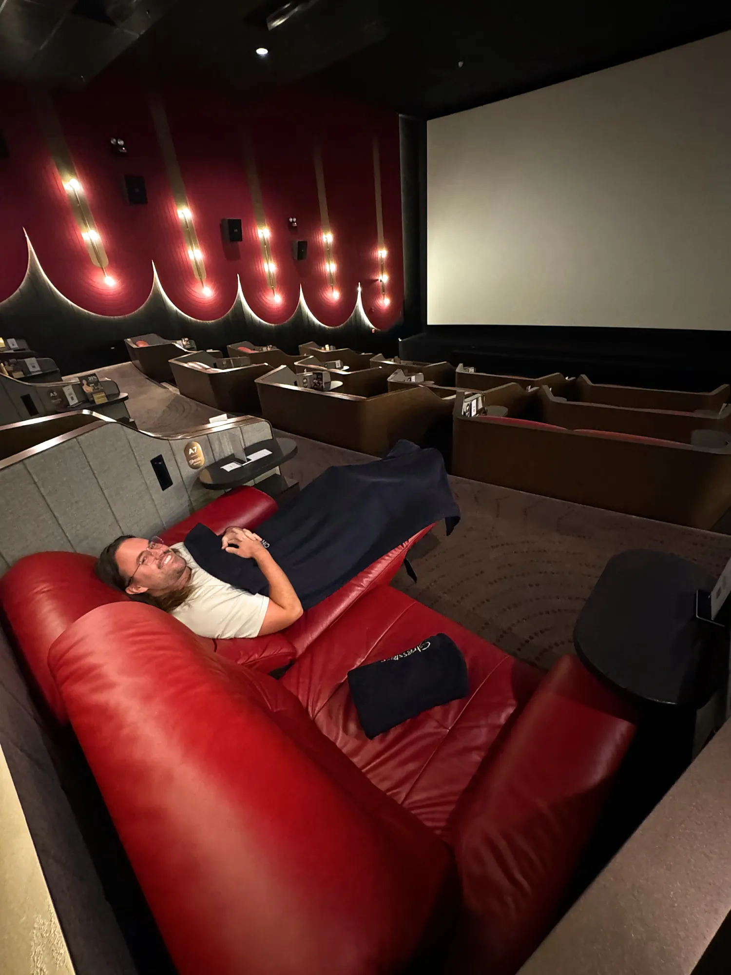 Cream lying back in a fully reclined red leather recliner facing the screen in a Maybank Comfort Cabin at Aurum Theatre, Kuala Lumpur.
