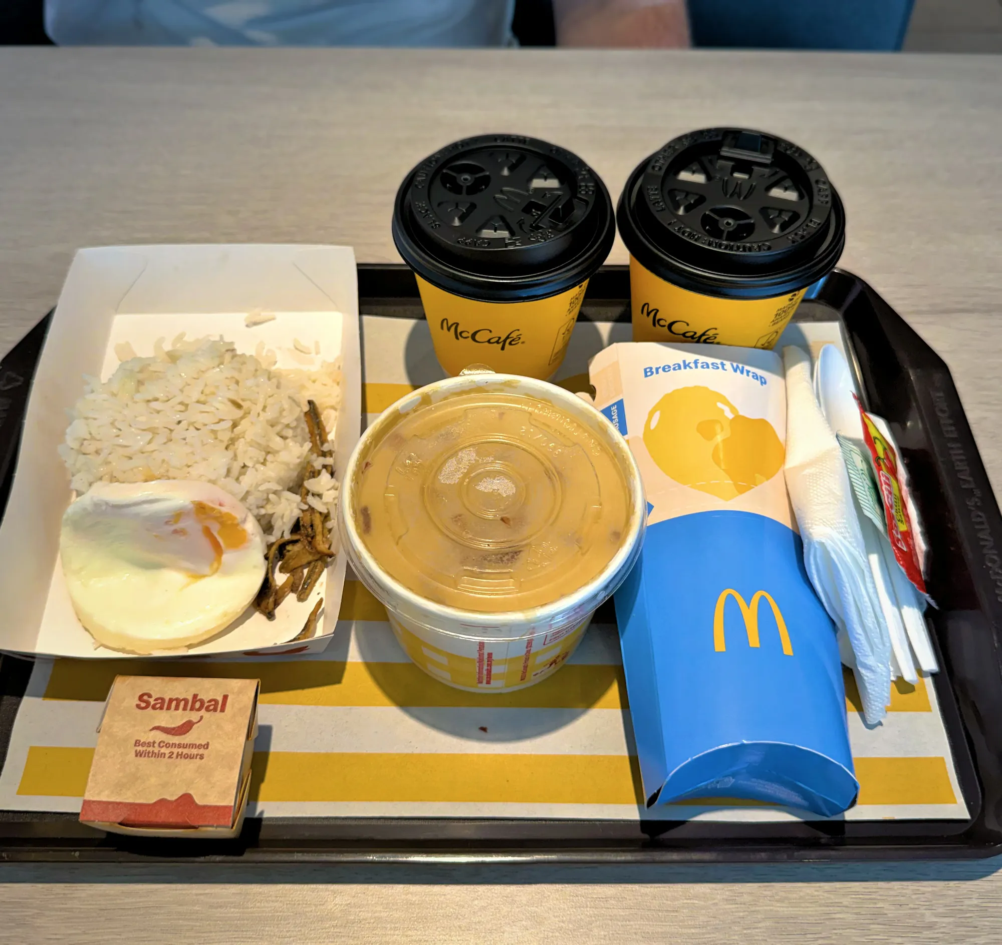 A full McDonald's Malaysian breakfast tray featuring Nasi Lemak McD with fried egg and sambal, chicken porridge, a breakfast wrap, and two hot McCafe drinks.