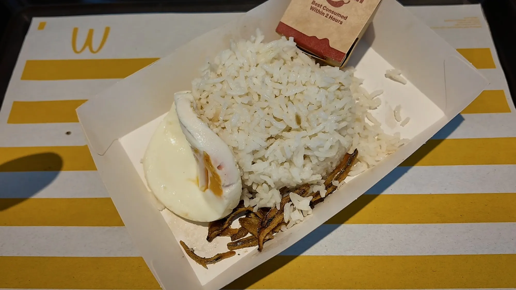 An opened Nasi Lemak McD box: coconut rice, a fried egg, and fried anchovies, with the sambal packet behind