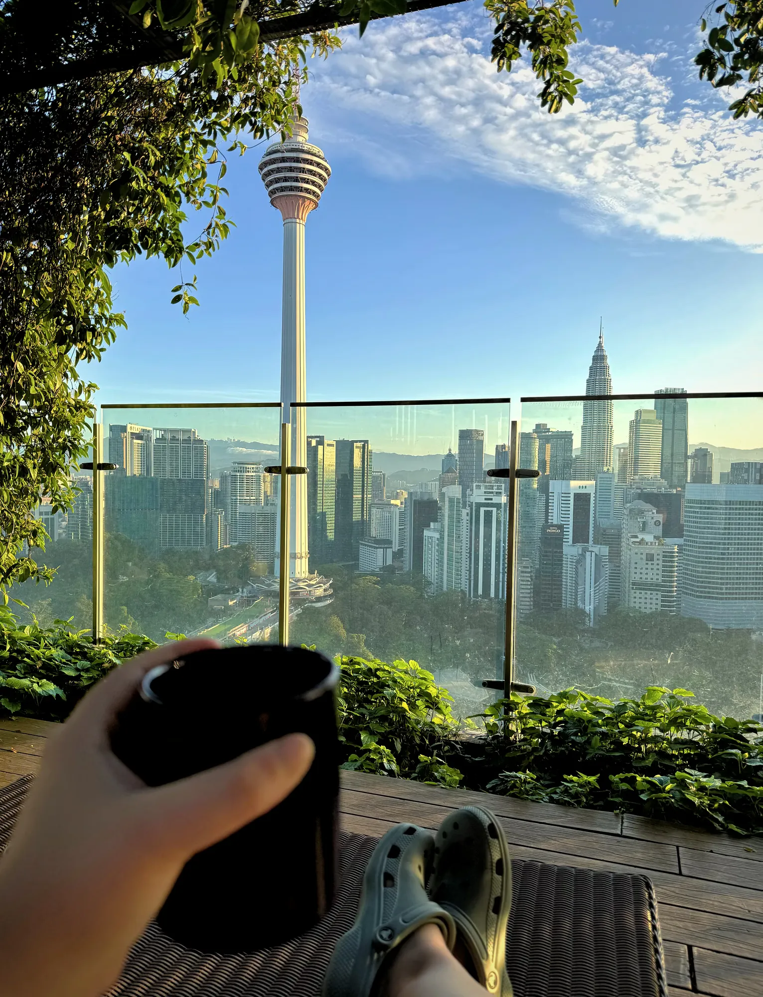 A hand holding a coffee mug on the sunny rooftop deck at Ceylonz Suites, with the KL Tower ahead.
