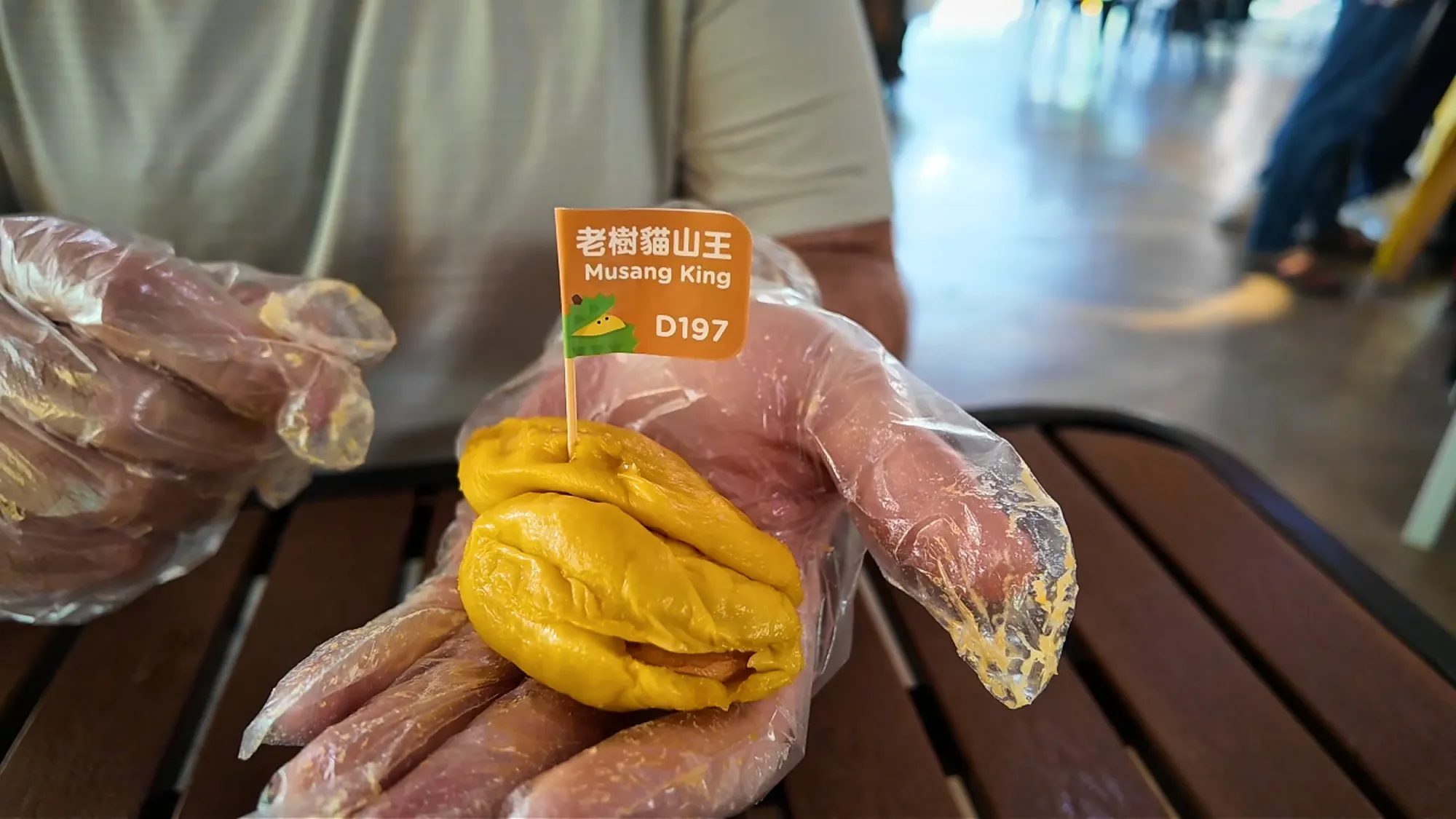 A gloved hand holding a rich golden Musang King durian piece with an orange flag marked D197.