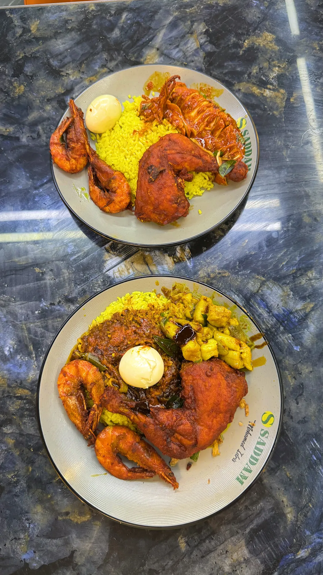 Two plates of nasi kandar at Saddam with fried chicken, prawns, squid, and boiled eggs on a dark marble table