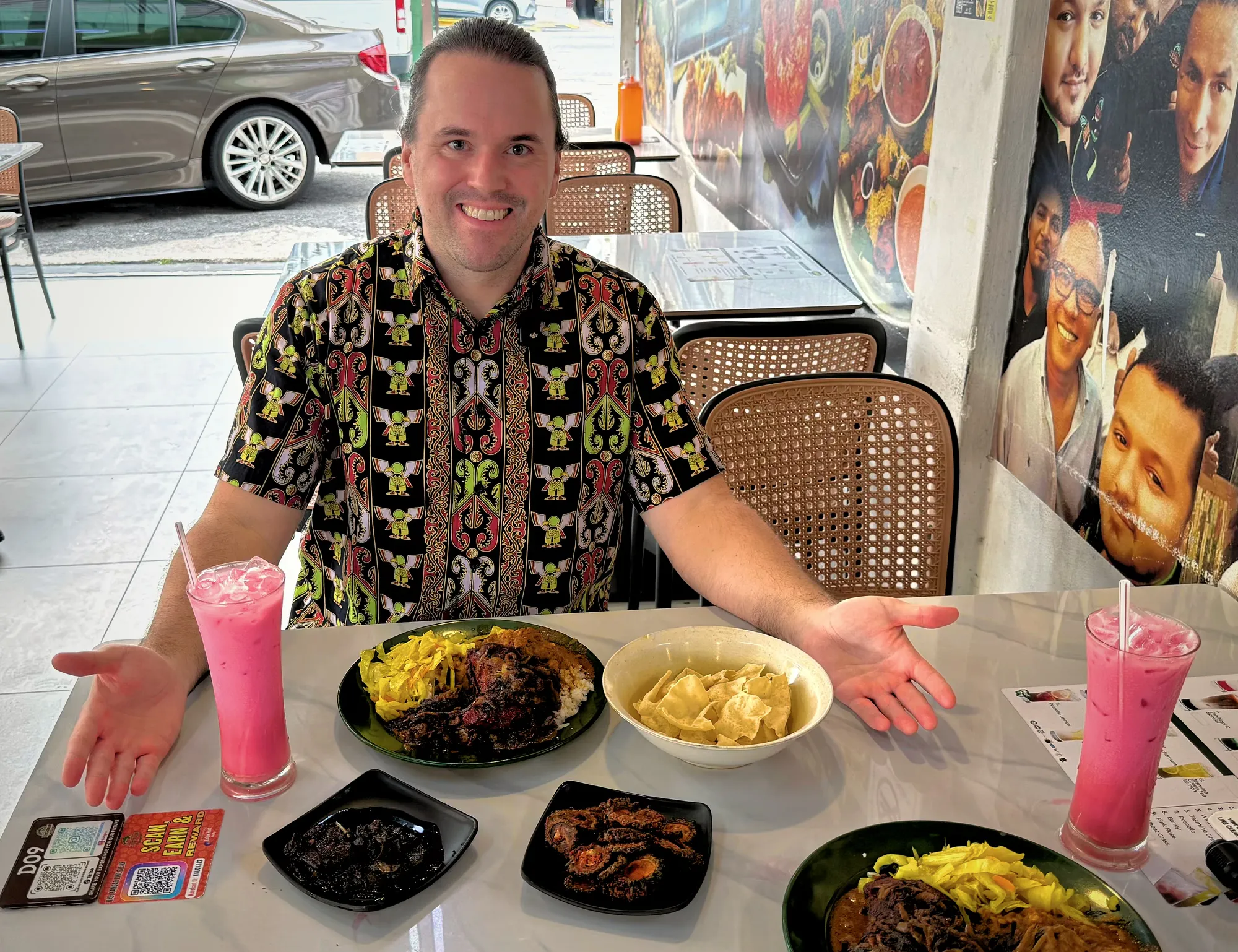 Arms open at a table full of nasi kandar plates, fried bitter gourd, keropok crackers, and pink sirap bandung drinks in Kuala Lumpur