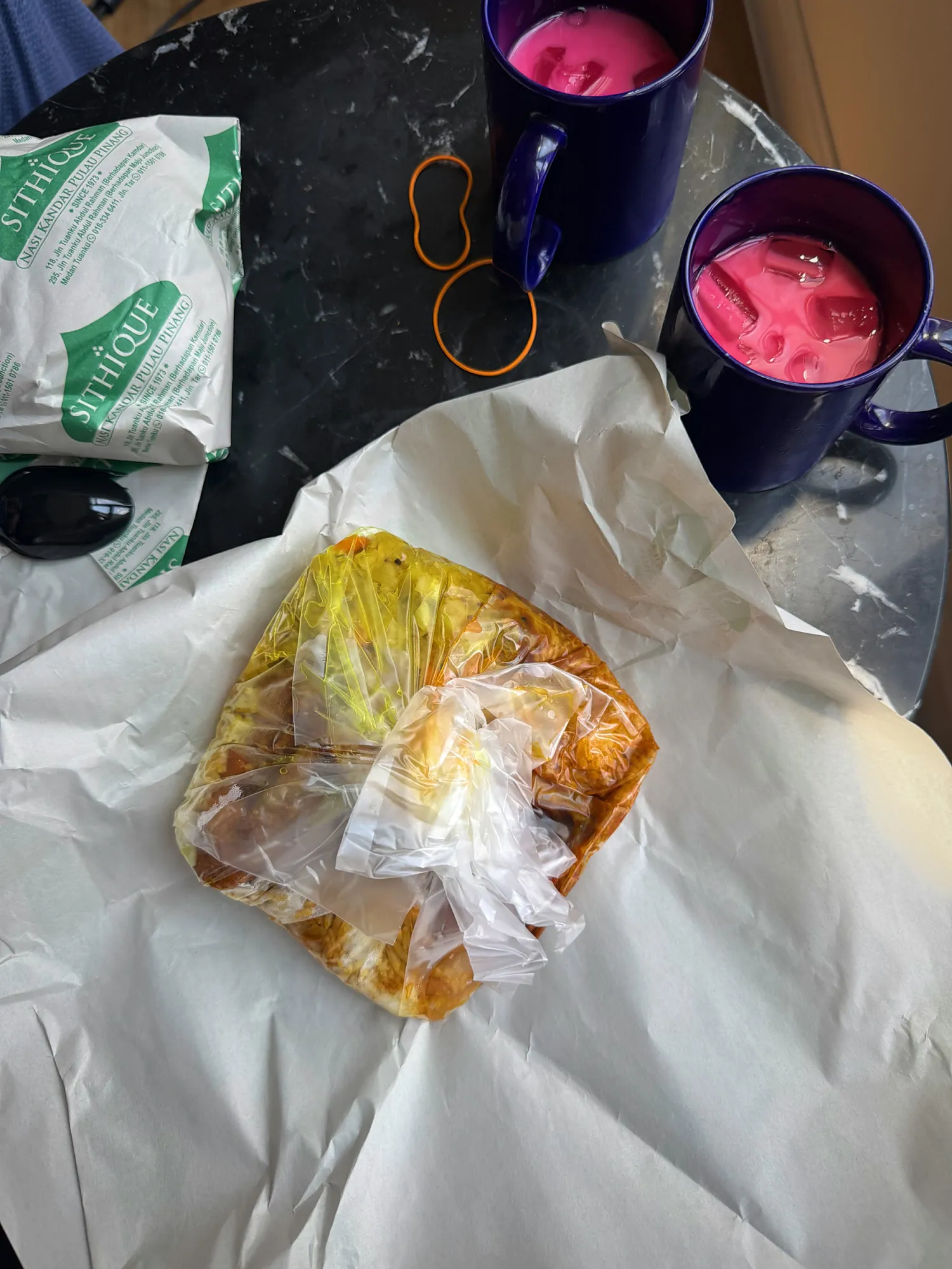 A takeaway nasi kandar in its paper wrapping next to two mugs of pink iced drinks on a table in Kuala Lumpur.