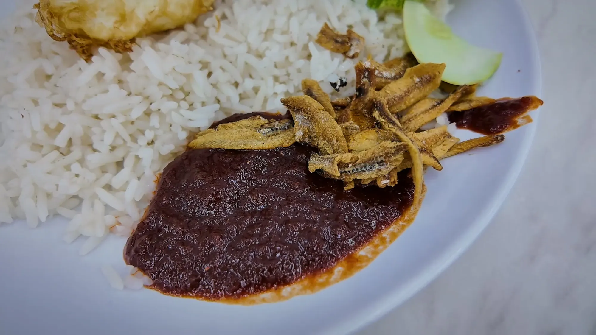 Close-up of Wanjo's dark sambal with fried anchovies, coconut rice, and cucumber on a white plate.