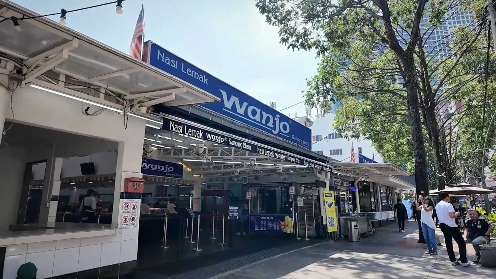 The storefront of Nasi Lemak Wanjo in Kampung Baru, Kuala Lumpur, with its big blue sign, under a clear morning sky.