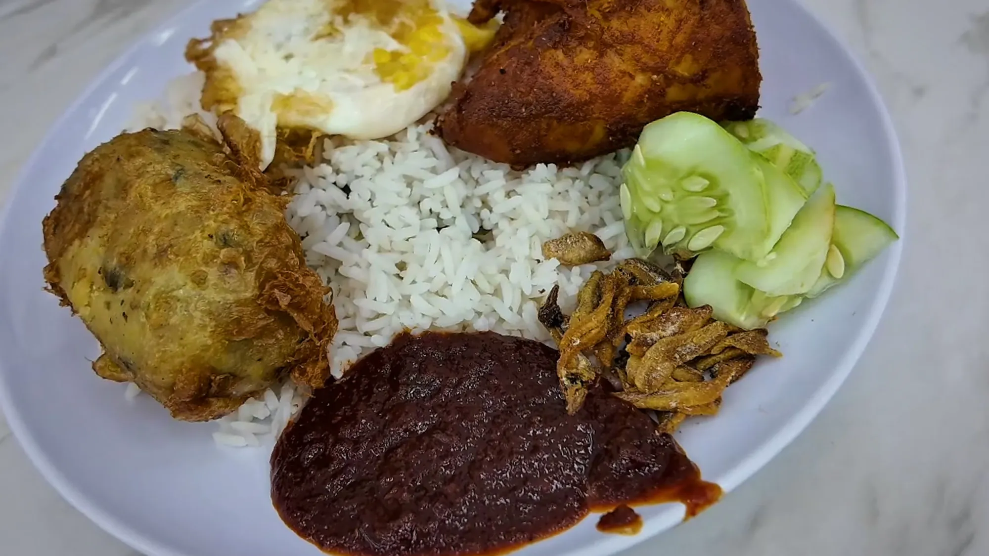 A begedil potato cake next to fried chicken, a fried egg, cucumber, anchovies, and dark sambal on a plate of nasi lemak at Wanjo.