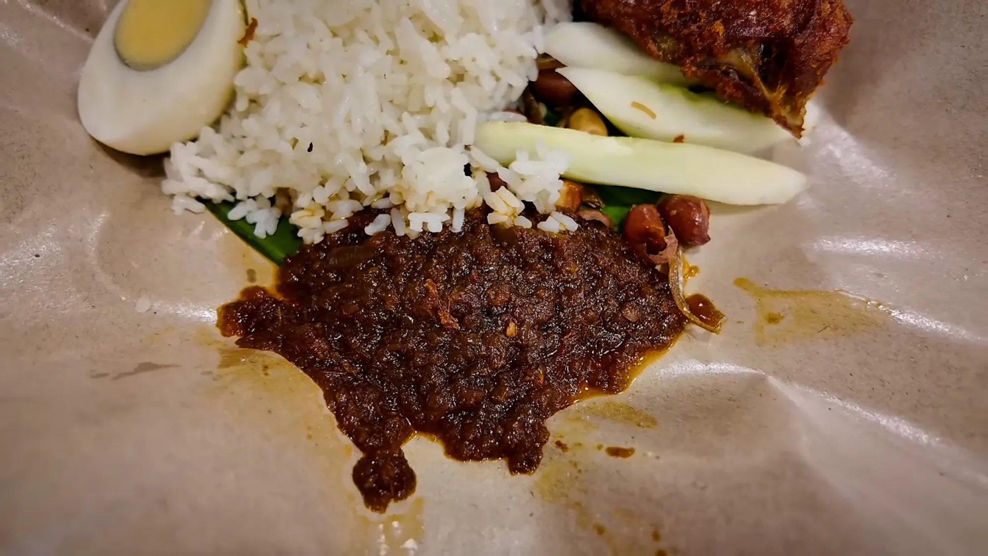 Close-up of a serving of nasi lemak with rice, hard-boiled egg, dark sambal, cucumber slices, and fried chicken.