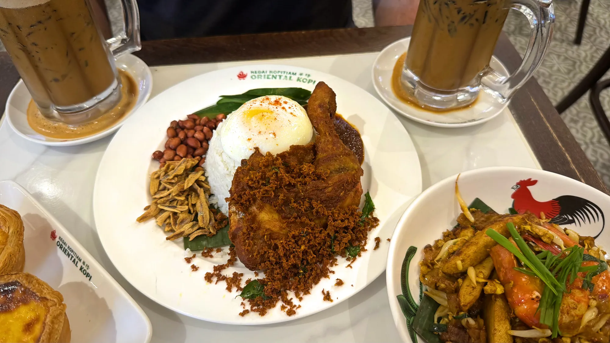 Close-up of Oriental Kopi nasi lemak with fried chicken, sambal, cucumber, and peanuts on a branded plate.