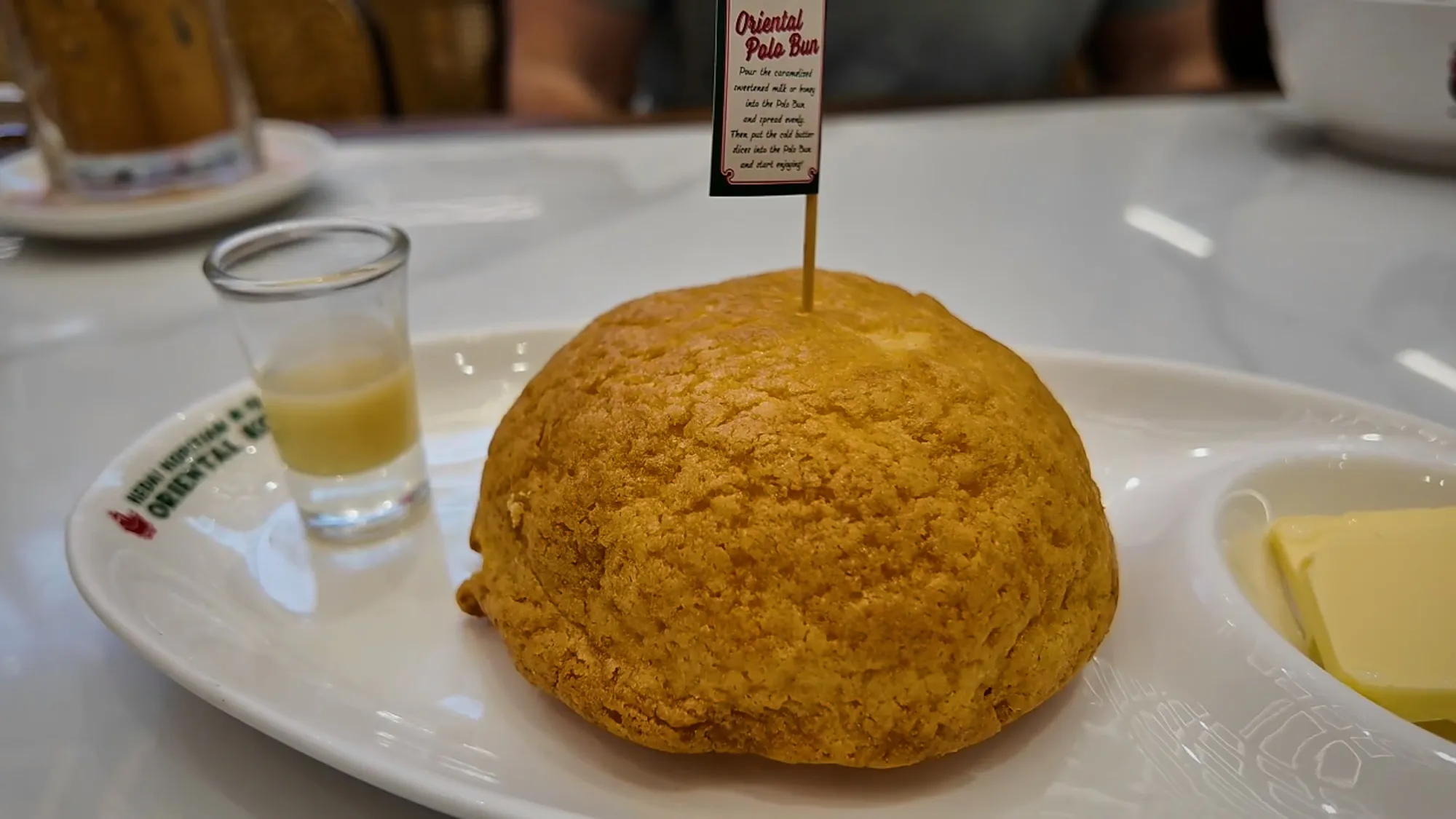 An Oriental Kopi polo bun on a plate with a small glass of sweetened milk and cold butter slices.