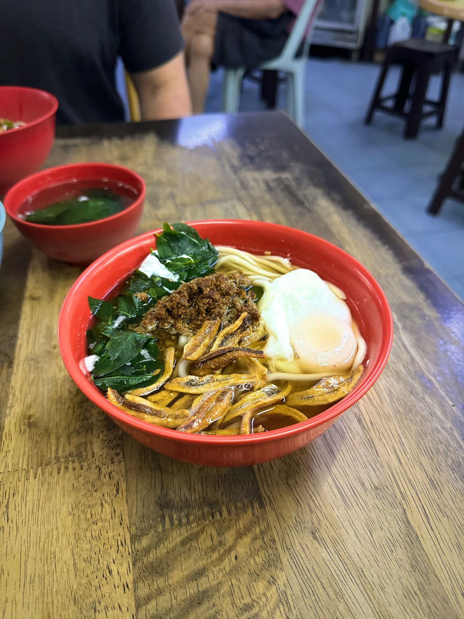 A red bowl of soup chili pan mee with leafy greens, a poached egg, minced pork, and anchovies, with a small bowl of clear soup beside it at Kin Kin.