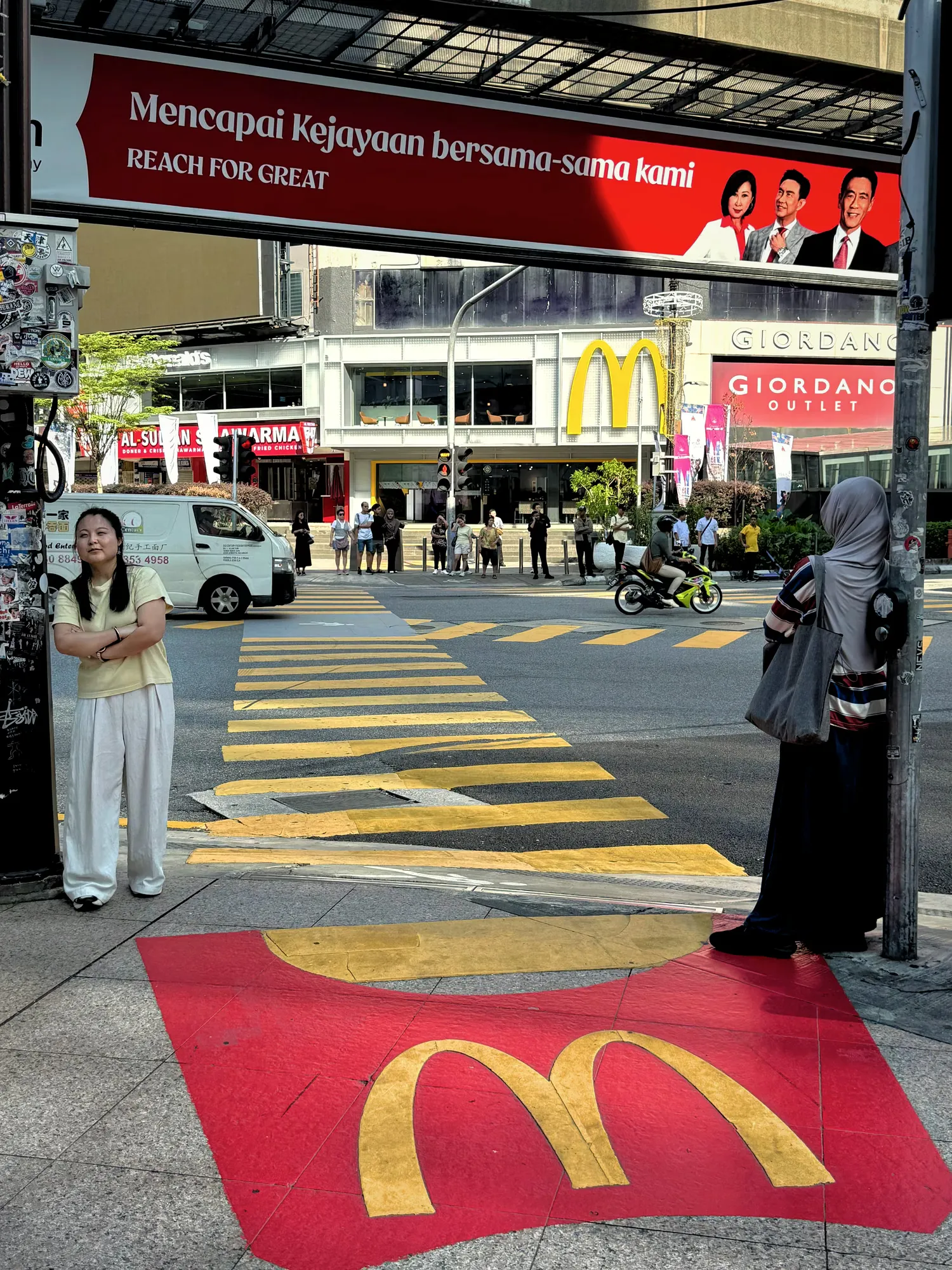 Two women waiting at a pedestrian crossing in Bukit Bintang, Kuala Lumpur, with a McDonald's fry carton painted on the pavement.