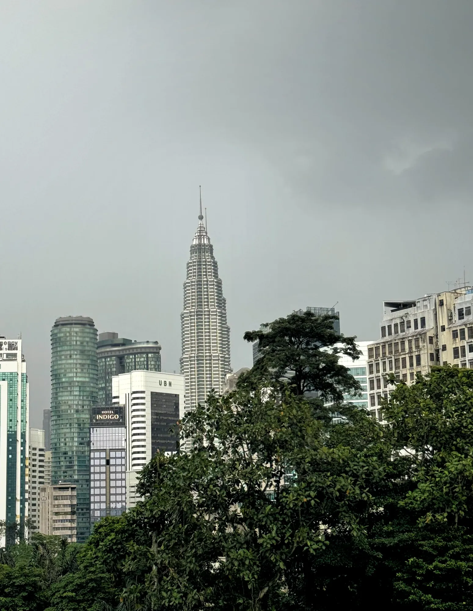 The silver spire of a Petronas Twin Tower rising above office towers and green treetops under a gray sky in Kuala Lumpur.