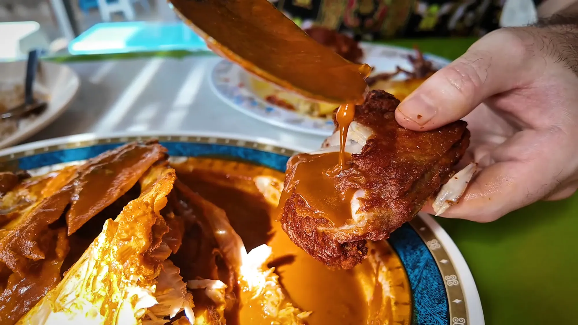 A spoon pouring fish head curry over a piece of fried chicken beside the fish head platter in Kuala Lumpur.