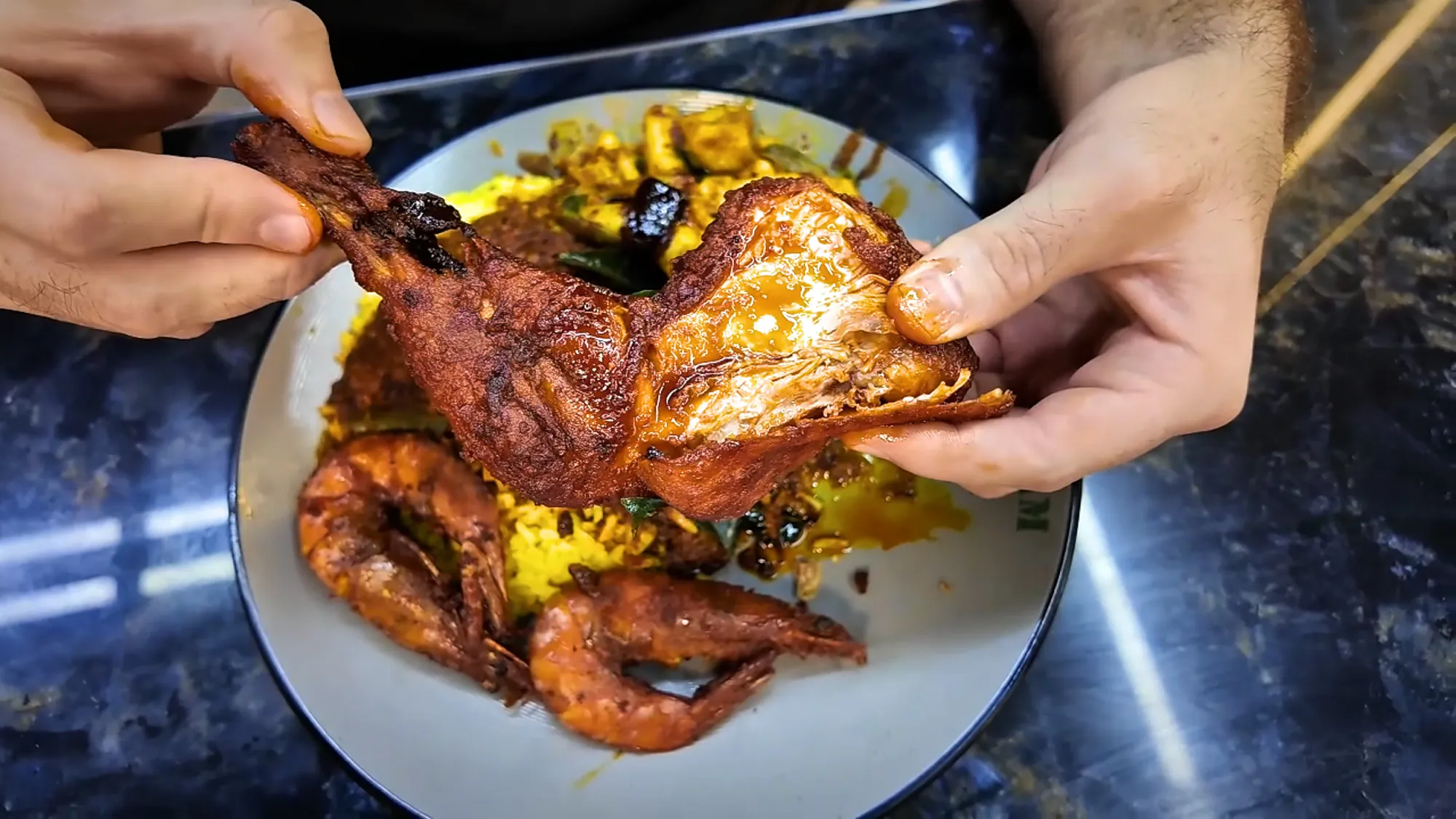 Hands pulling apart a spiced fried chicken leg over yellow rice and prawns at Saddam Nasi Kandar in Kuala Lumpur.