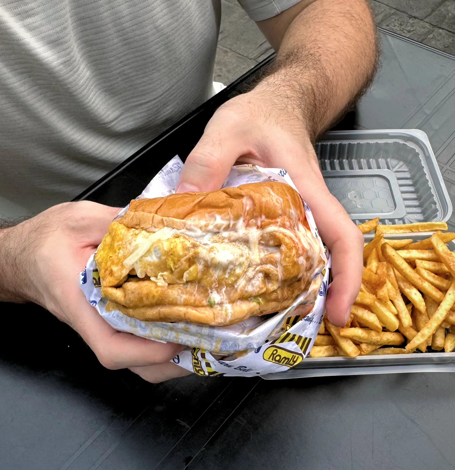 A Ramly Special burger wrapped in a thin egg and covered in sauce, held over a tray of fries at an outdoor table on Jalan Alor, Kuala Lumpur