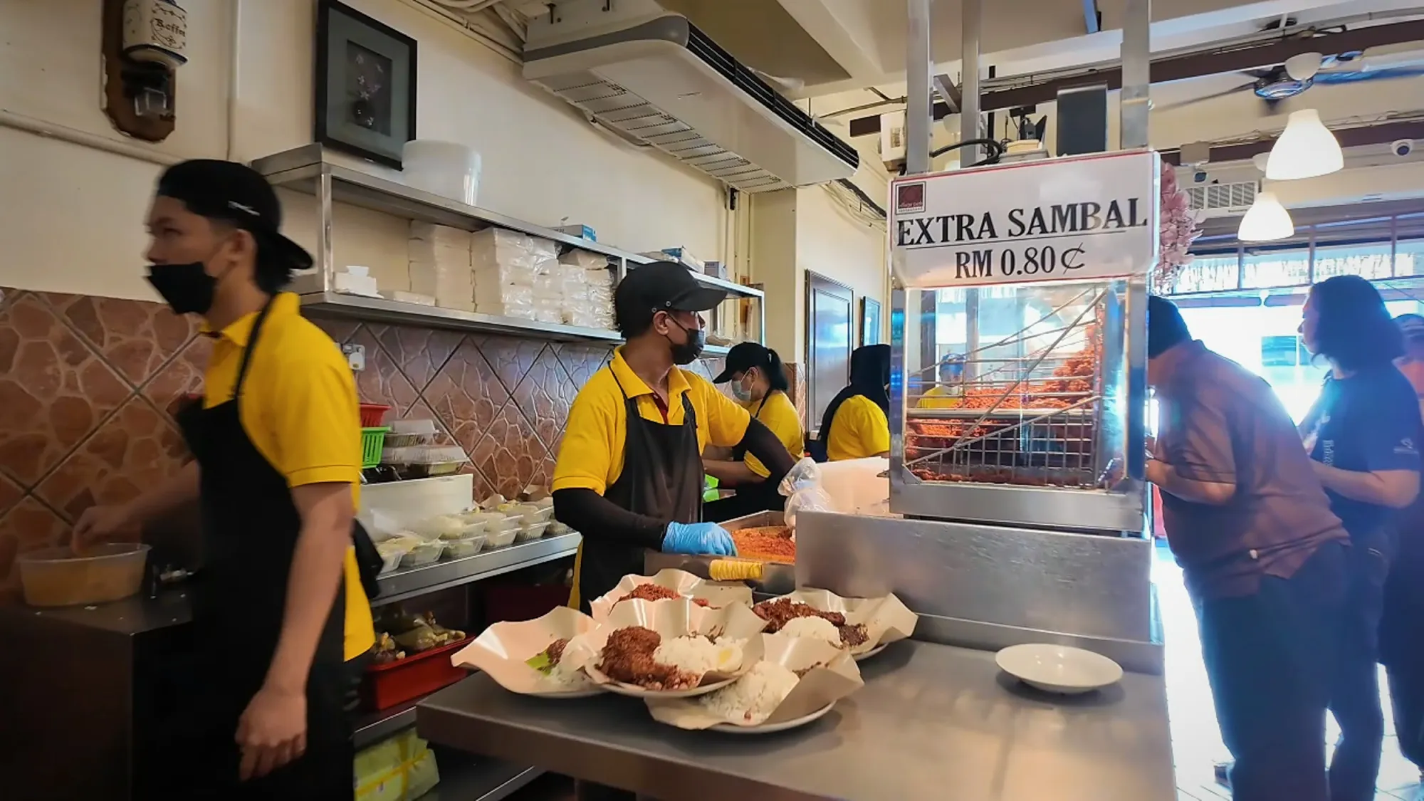 Staff in yellow shirts and black aprons preparing dishes at a counter in a busy restaurant.