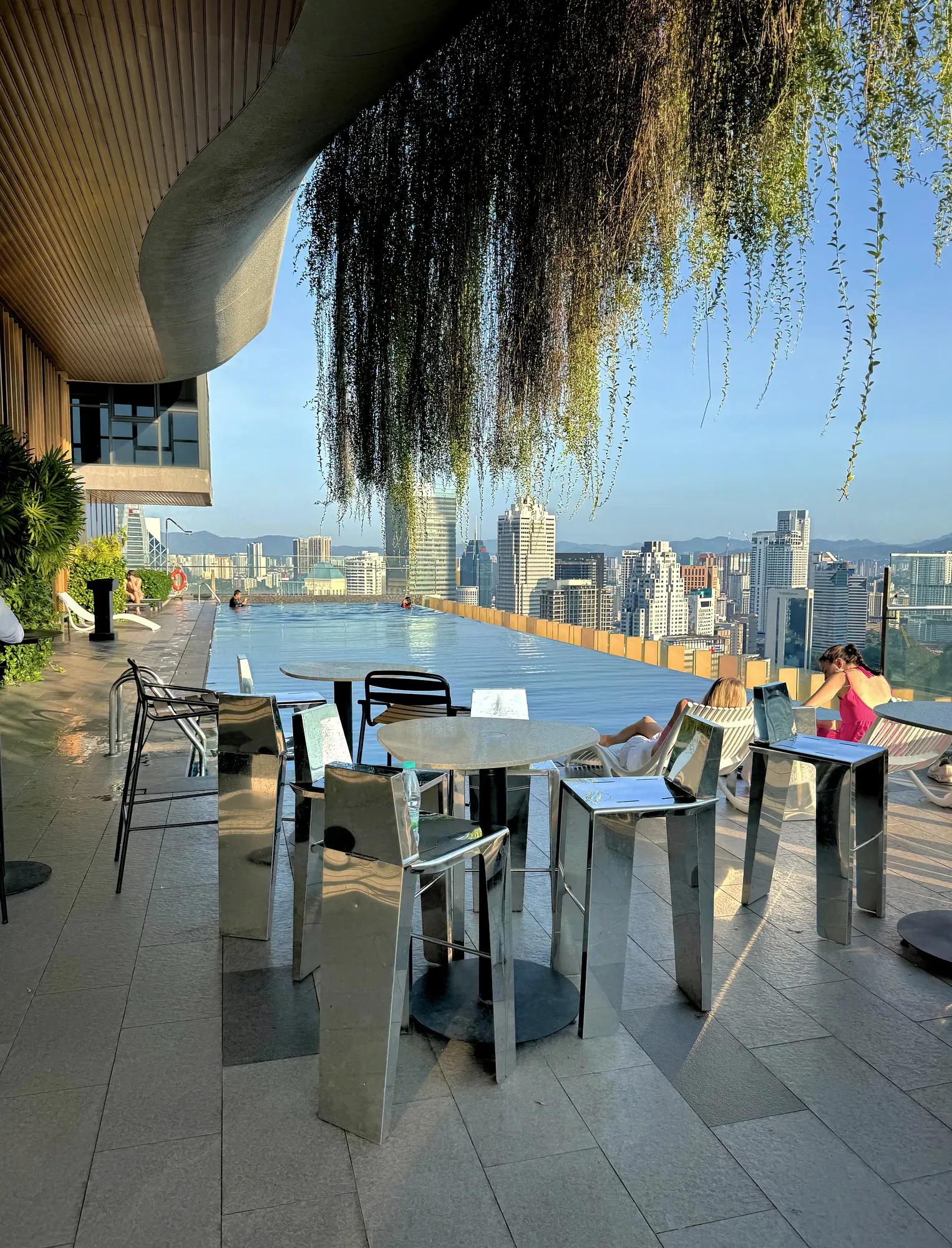 A rooftop infinity pool framed by metal bar stools and hanging vines, overlooking the Kuala Lumpur skyline on a clear morning.