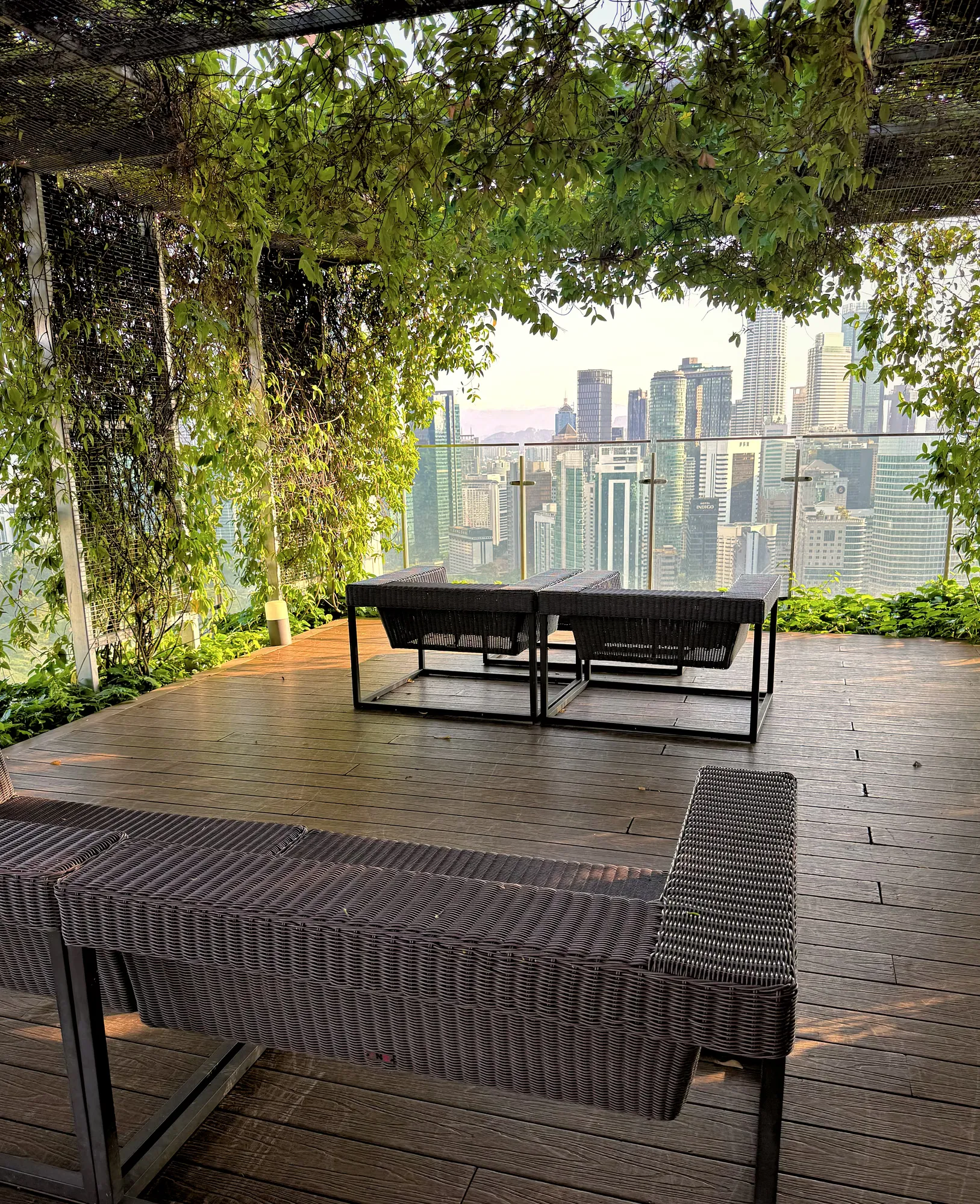 Woven patio sofas on a timber deck surrounded by climbing ivy, with Kuala Lumpur skyscrapers beyond, at Ceylonz Suites.
