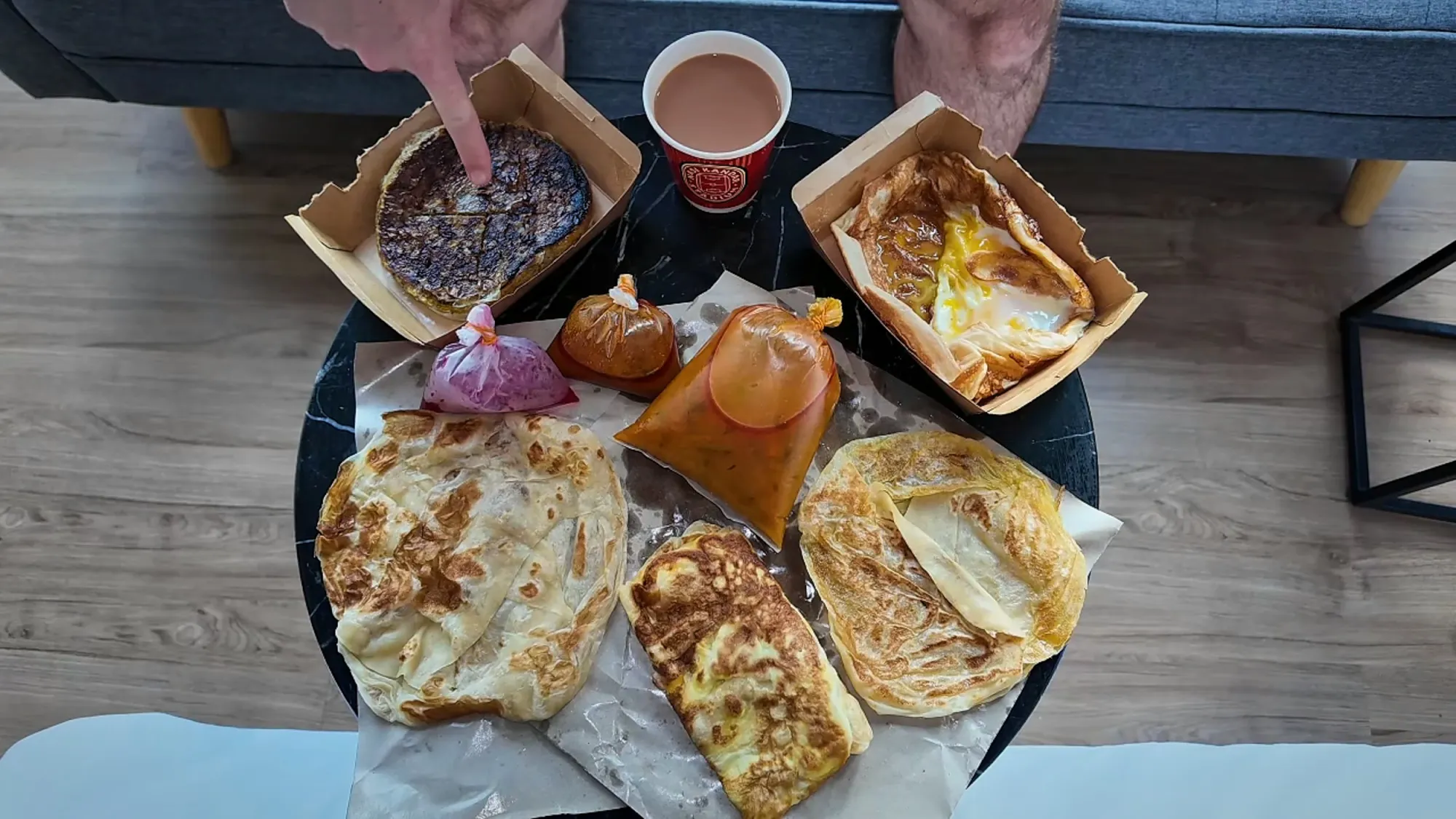 A spread of flaky roti canai, murtabak, and egg roti with takeaway bags of dhal and a hot teh tarik on a round black table in Kuala Lumpur
