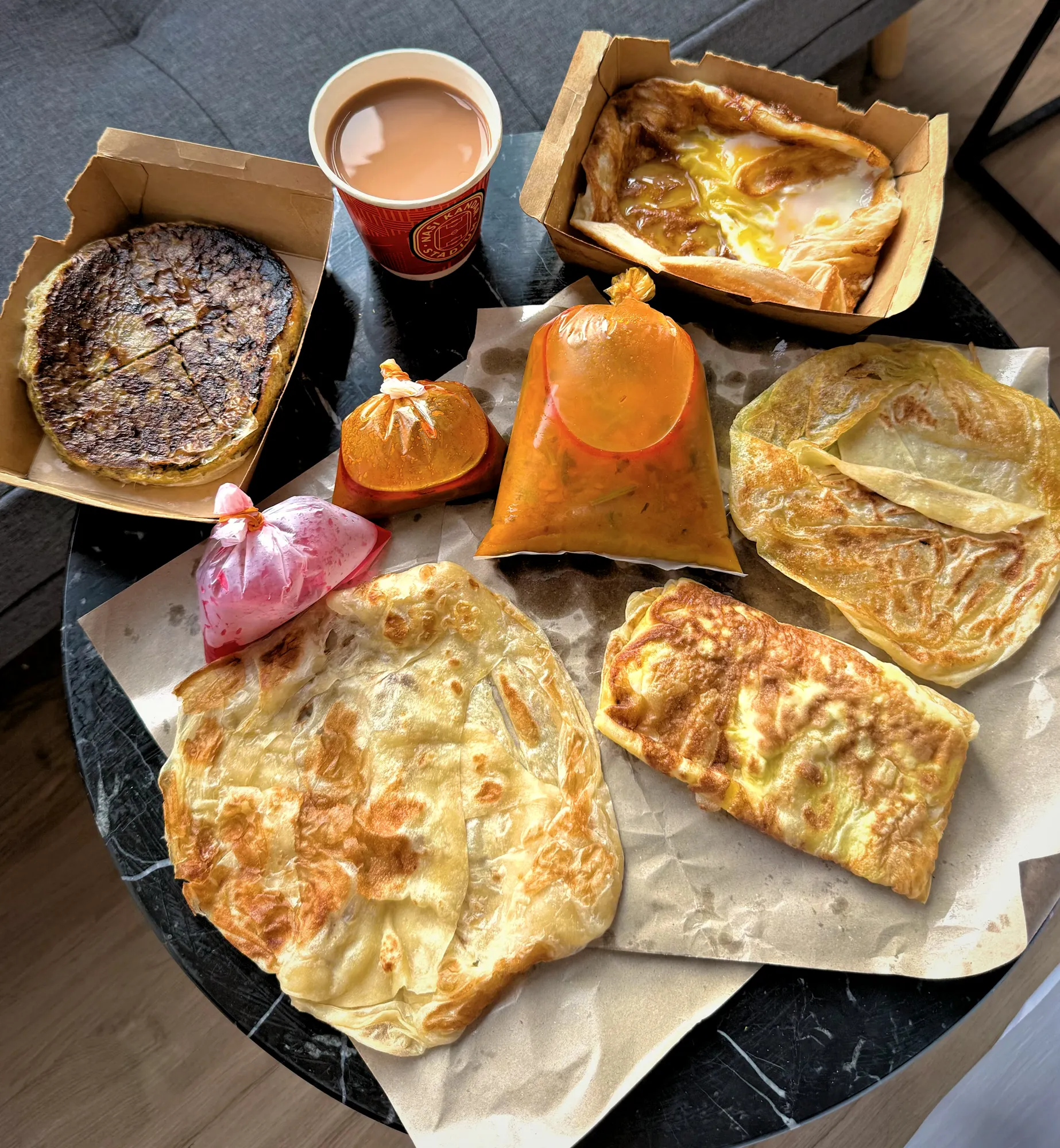 Flaky roti canai, murtabak, roti telur, and takeaway curry packets on a round black table beside hot teh tarik in Kuala Lumpur