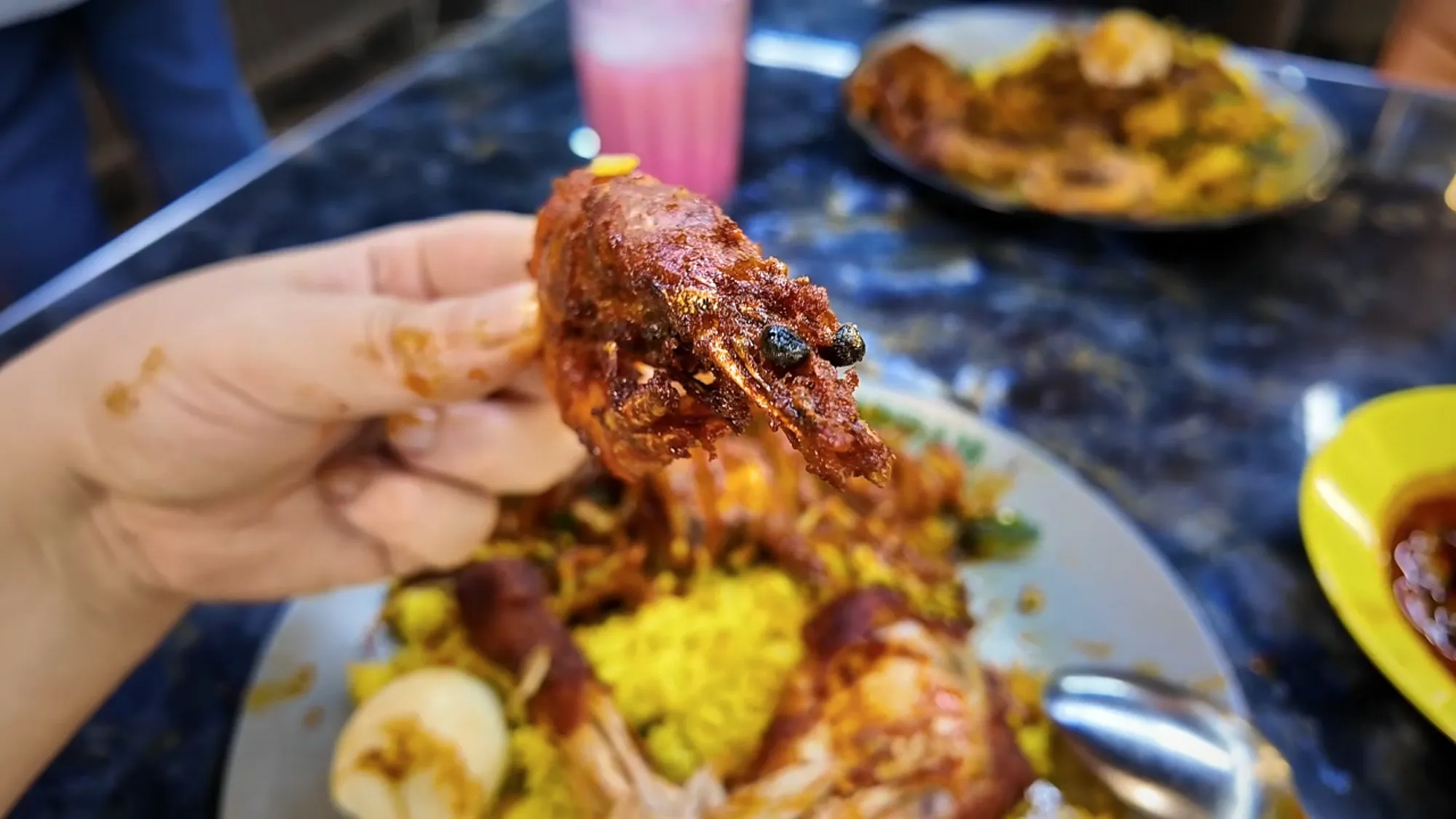 A hand holding up a big spiced prawn over a plate of yellow rice and curry at Saddam Nasi Kandar in Kuala Lumpur.