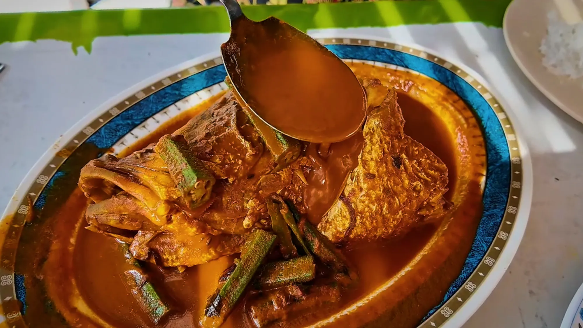 A spoon pouring red curry over a whole fish head with okra on a blue-rimmed platter at Anuar Curry Fish Head in Bangsar, Kuala Lumpur.