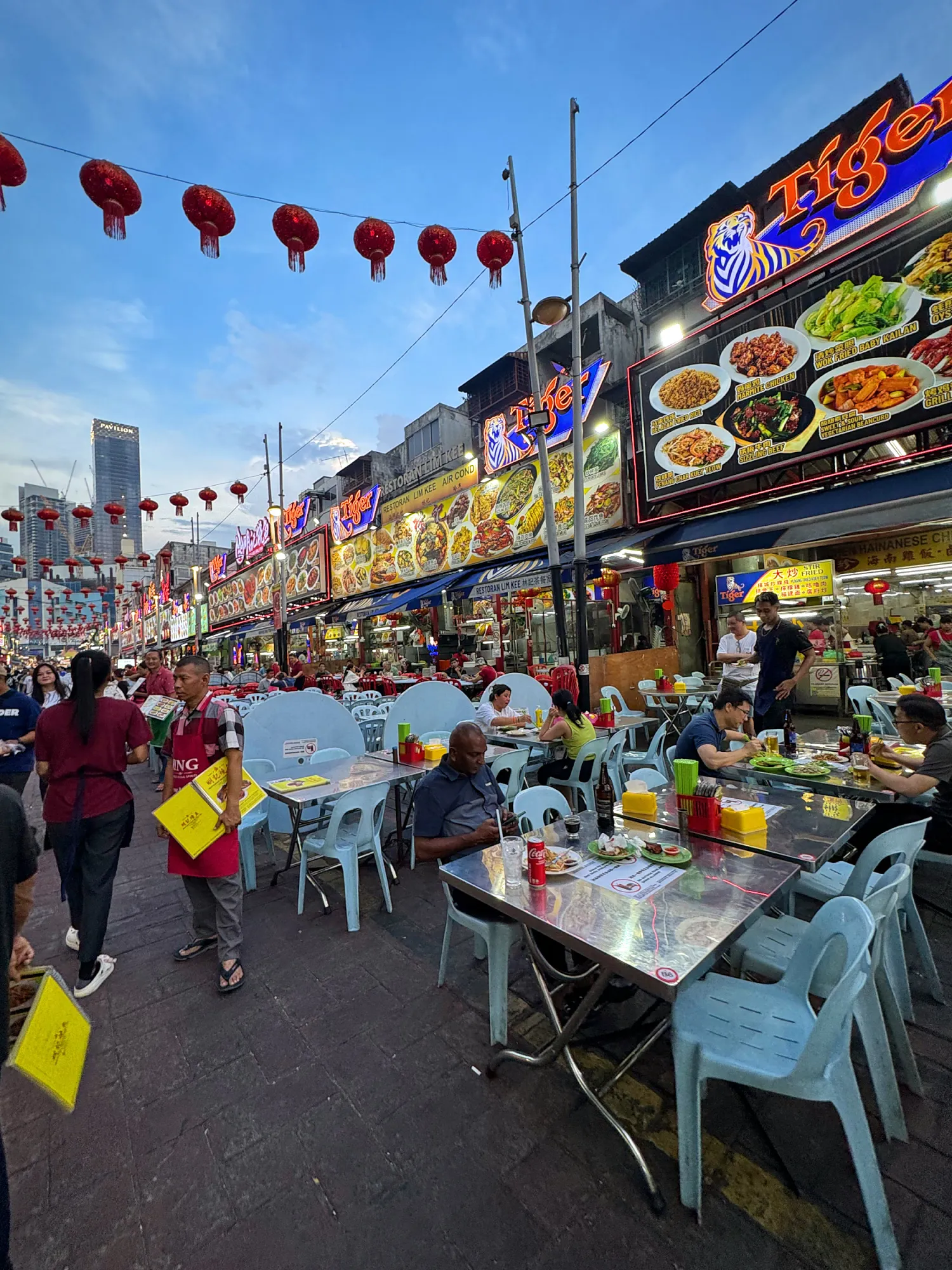 An outdoor street dining area with tables, diners, and bright neon restaurant signs under a twilight sky in Kuala Lumpur.