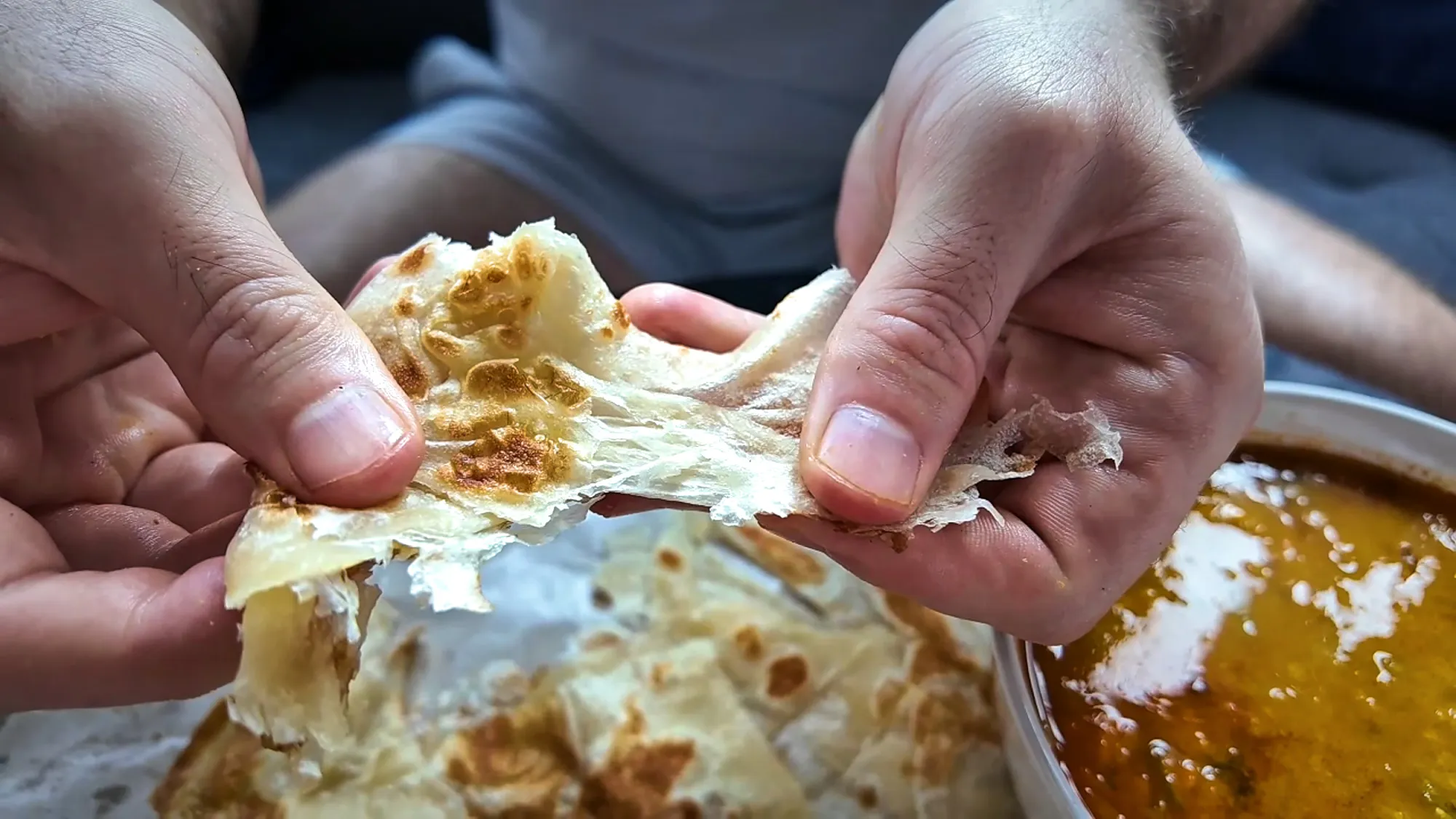 Hands tearing a piece of flaky roti canai apart to show its thin, stretchy layers, next to a bowl of dhal