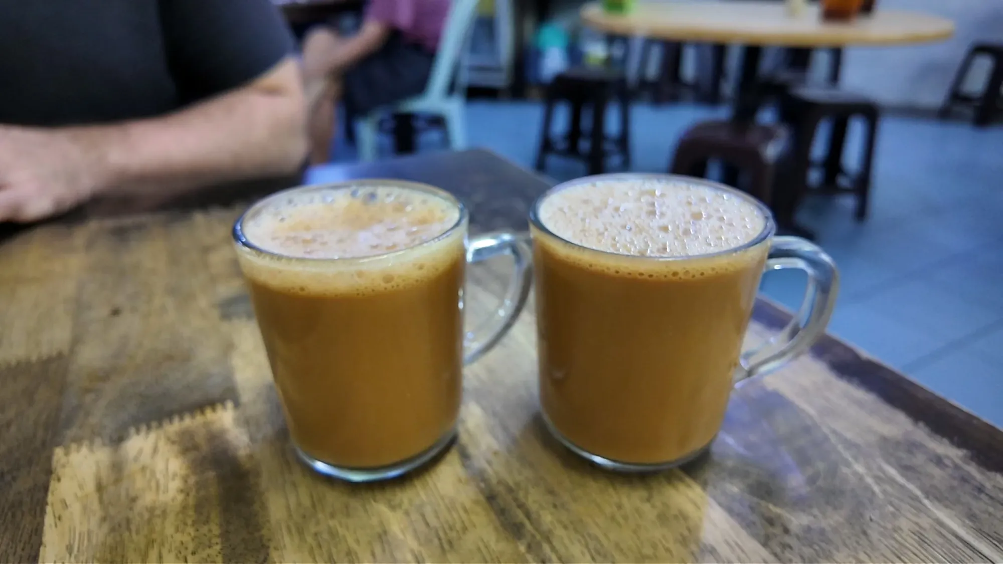Two glass mugs of frothy teh tarik on a wooden table at Restoran Kin Kin, Kuala Lumpur.
