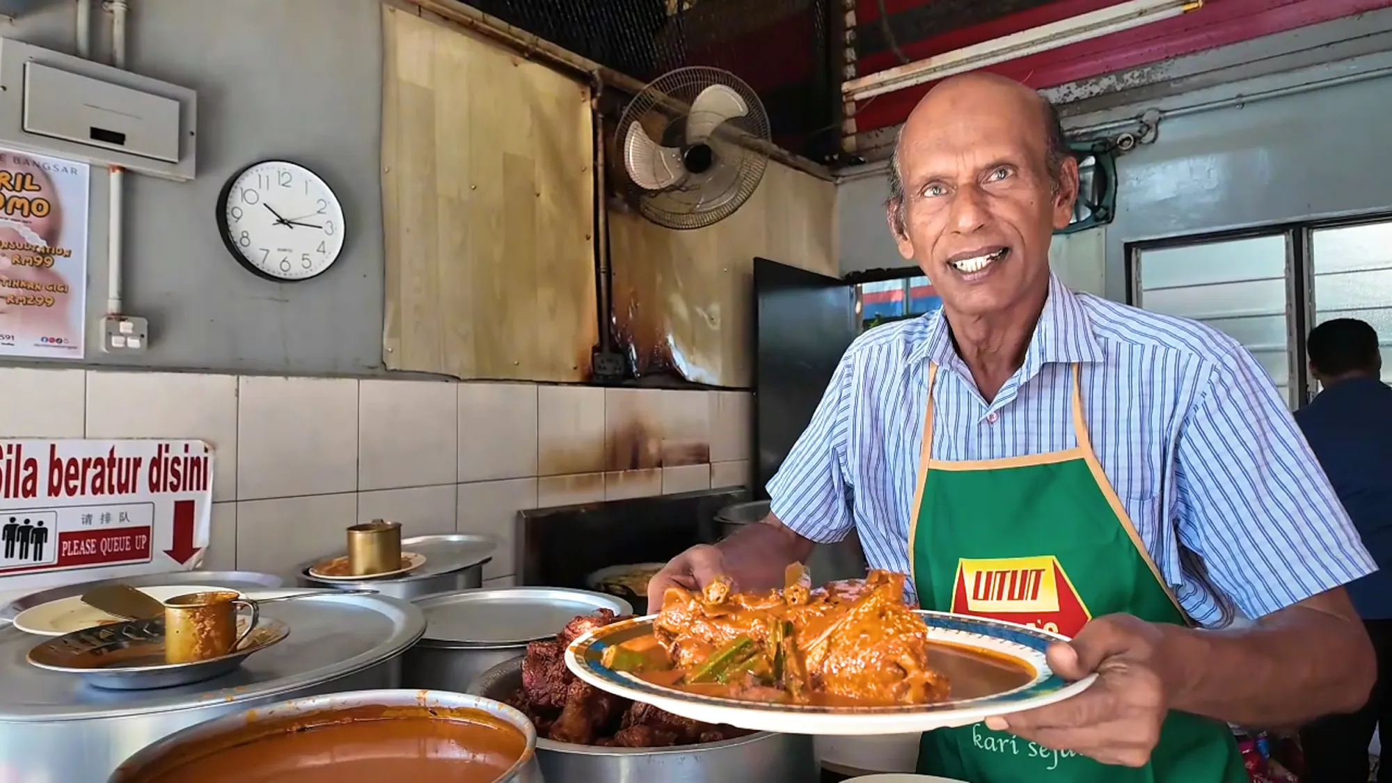 Anuar smiling as he holds out a platter of fish head curry with okra across the counter of his stall in Bangsar, Kuala Lumpur.