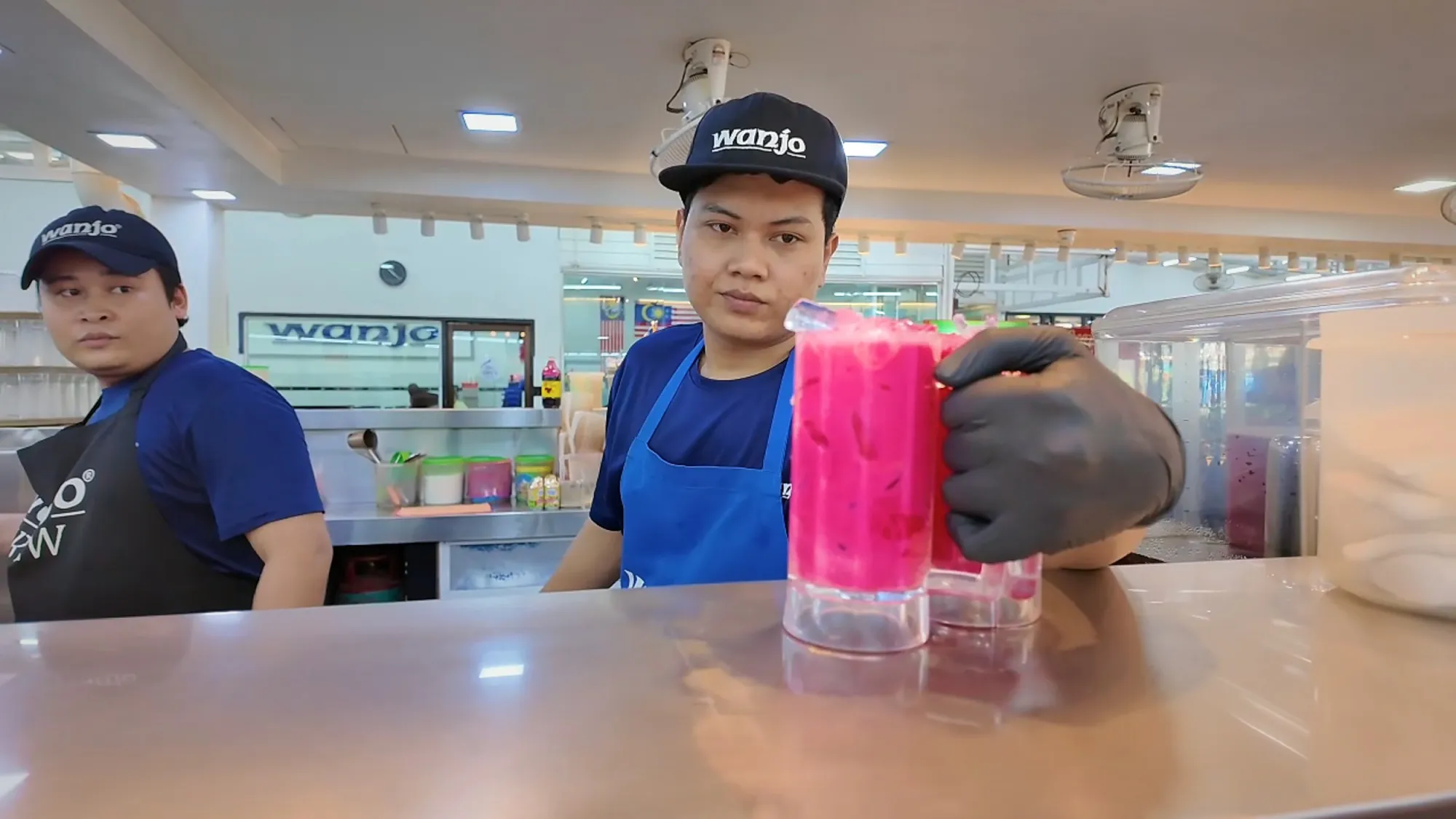 A Wanjo staff member in a blue apron and cap holding up two bright pink iced sirap bandung drinks across the counter.