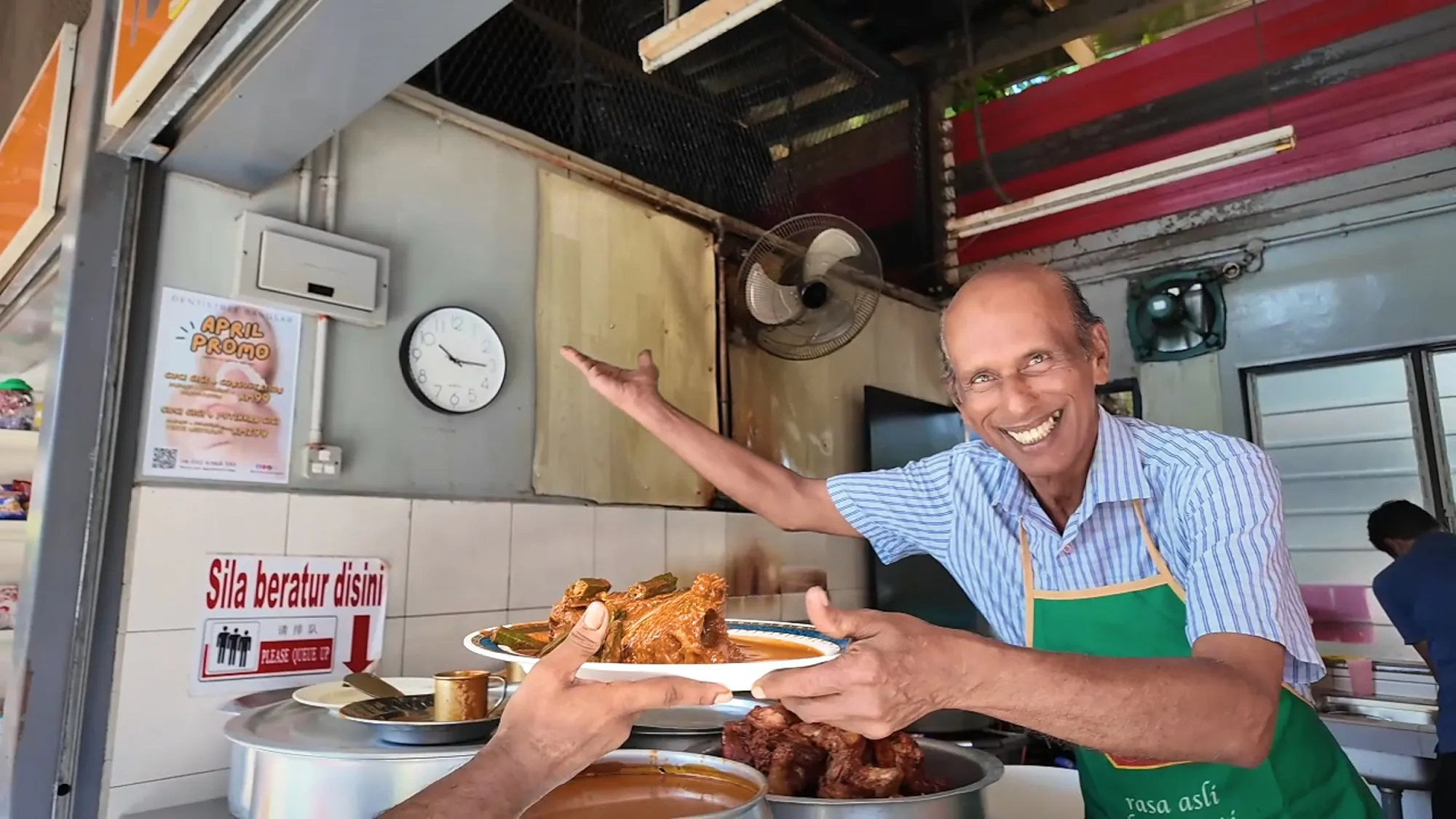 Anuar smiling as he holds out a platter of fish head curry with okra across the counter of his stall in Bangsar, Kuala Lumpur.