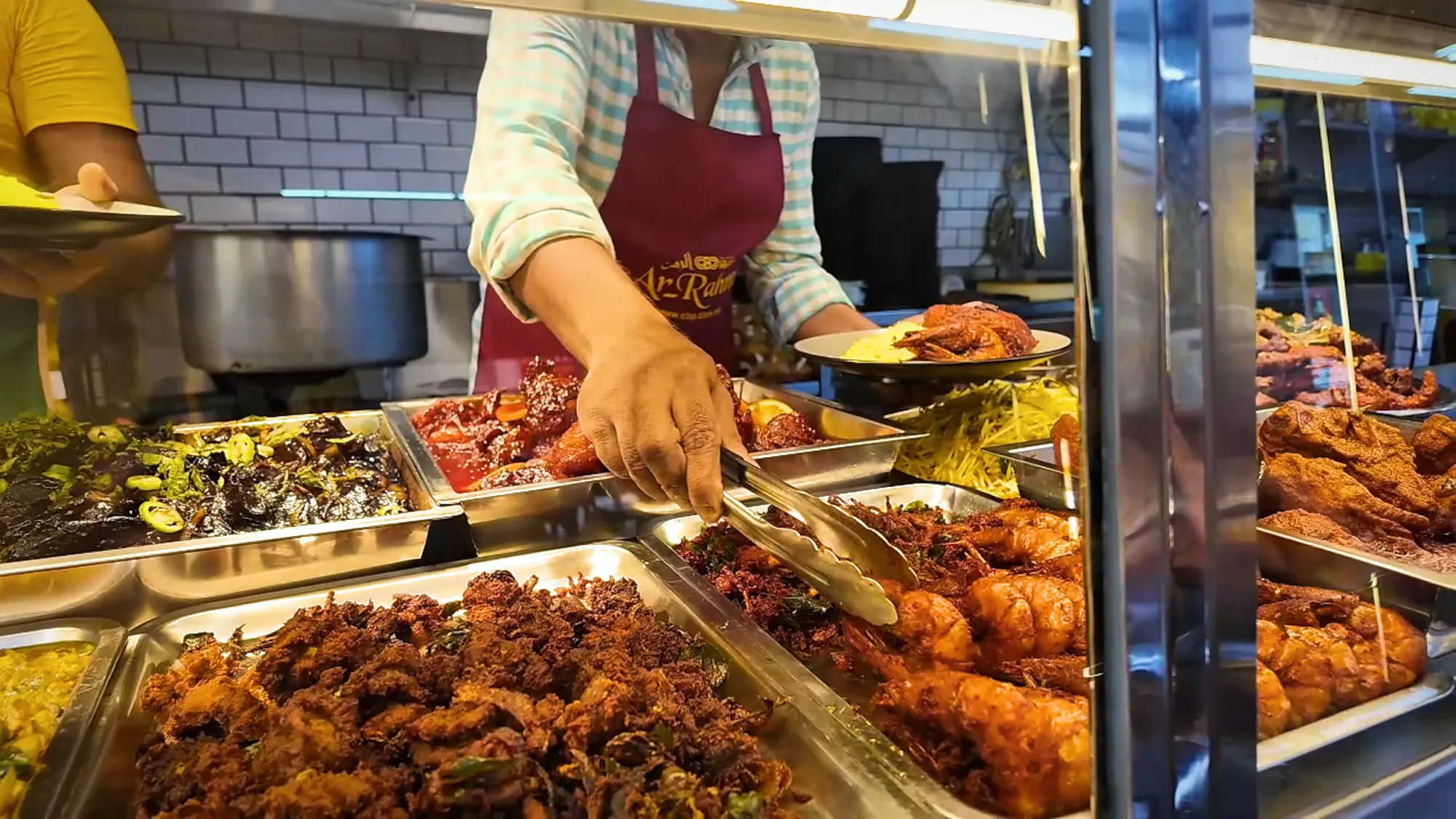A server using tongs to pick fried prawns and fried chicken from the counter trays at Saddam Nasi Kandar in Kuala Lumpur.